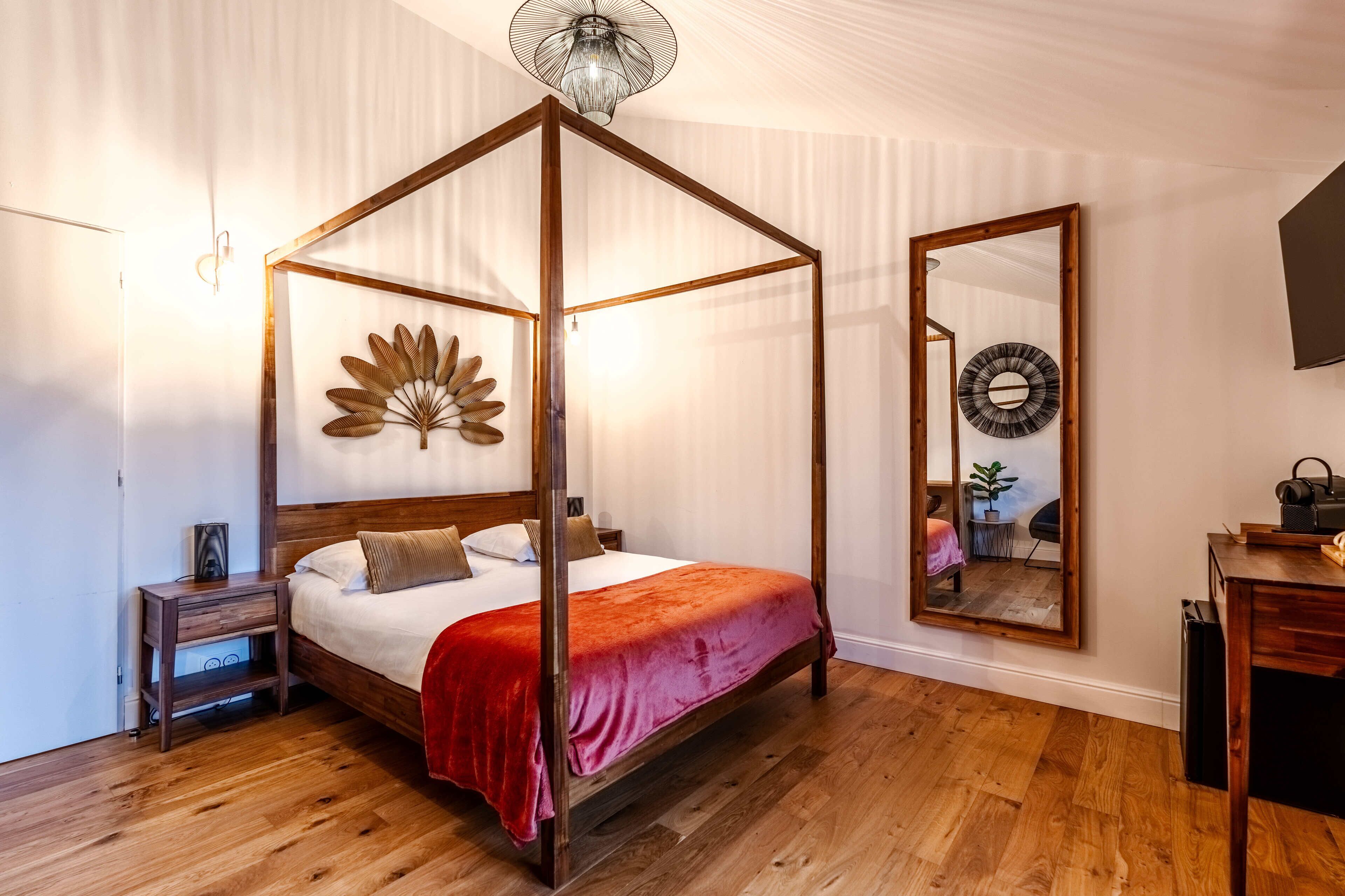 Una cama con dosel de madera y colcha roja, una mesita de noche de madera con lámpara, un espejo grande en la pared y suelo de madera.