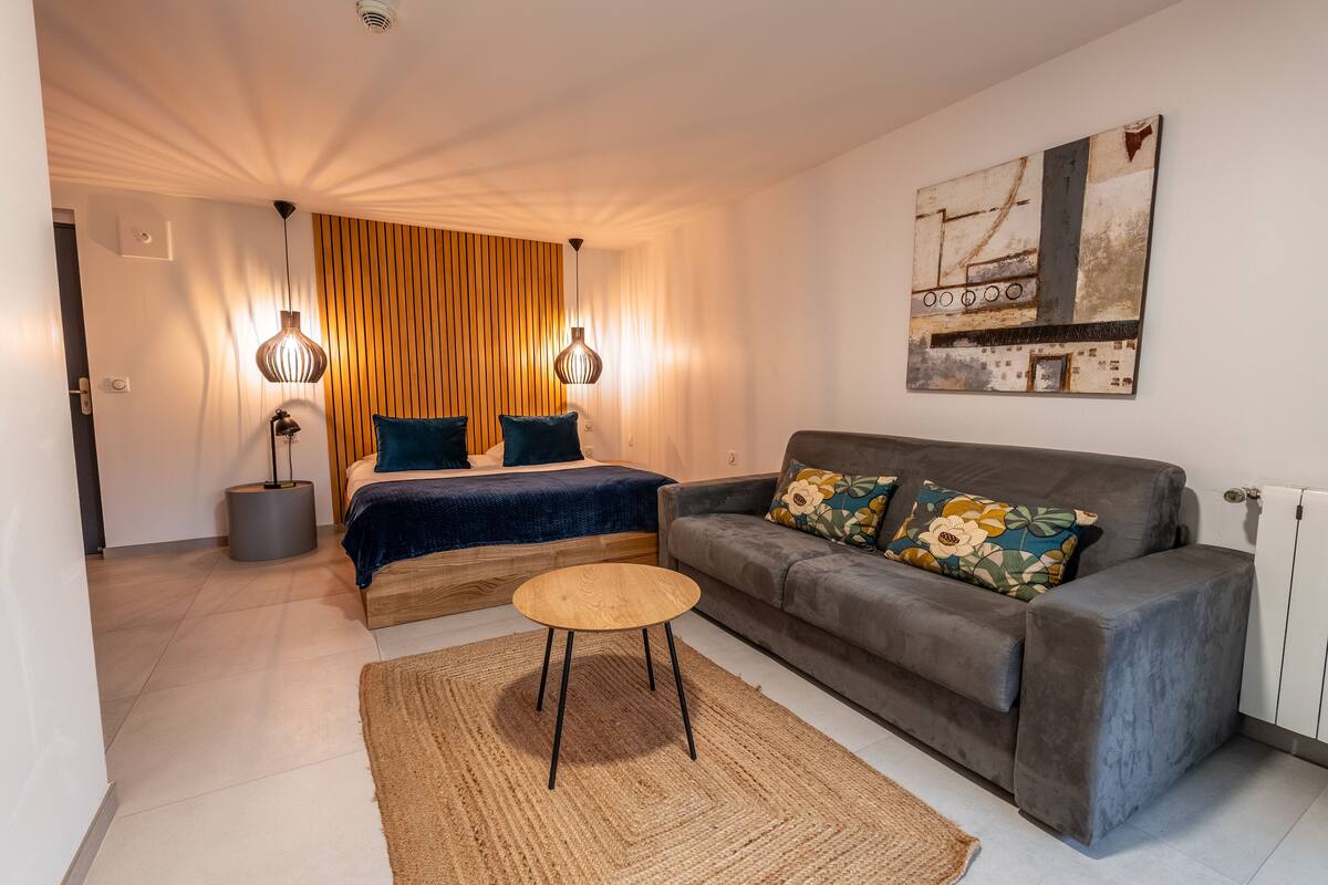A modern bedroom with a bed, a grey sofa, a small round table, and a large abstract painting on the wall.