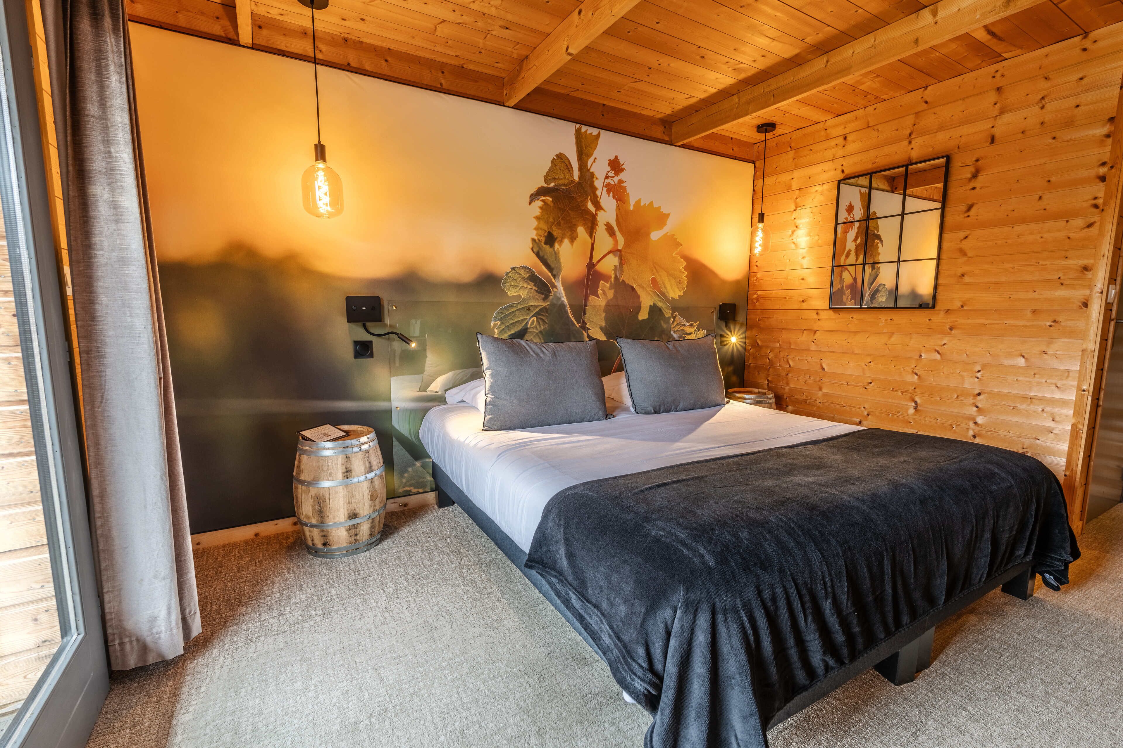 A wooden cabin bedroom with a bed, pillows, a nightstand, a barrel, and a wall-mounted light fixture.