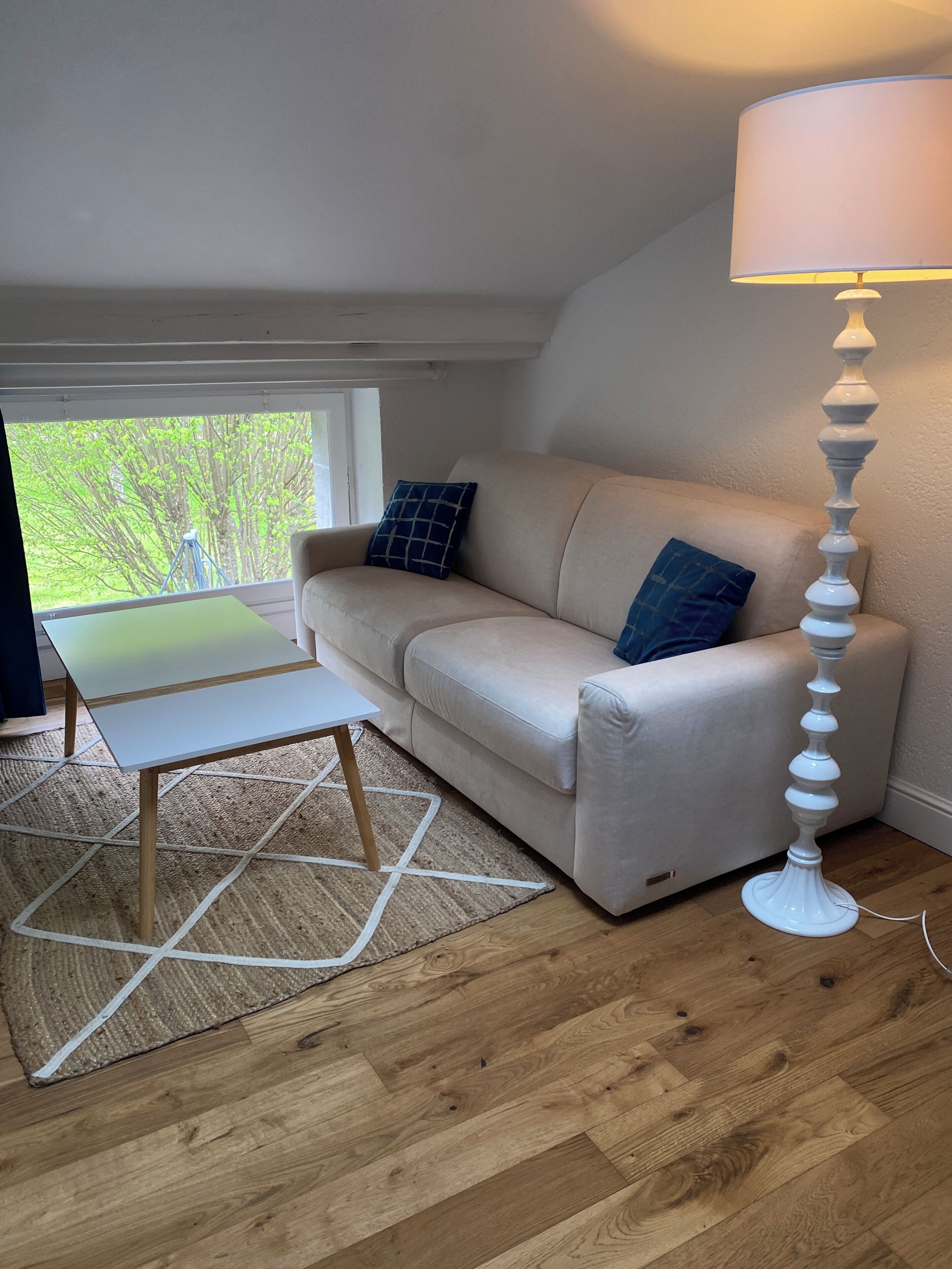 Een kamer met een bank, een klein tafeltje, een lamp en uitzicht door een raam.