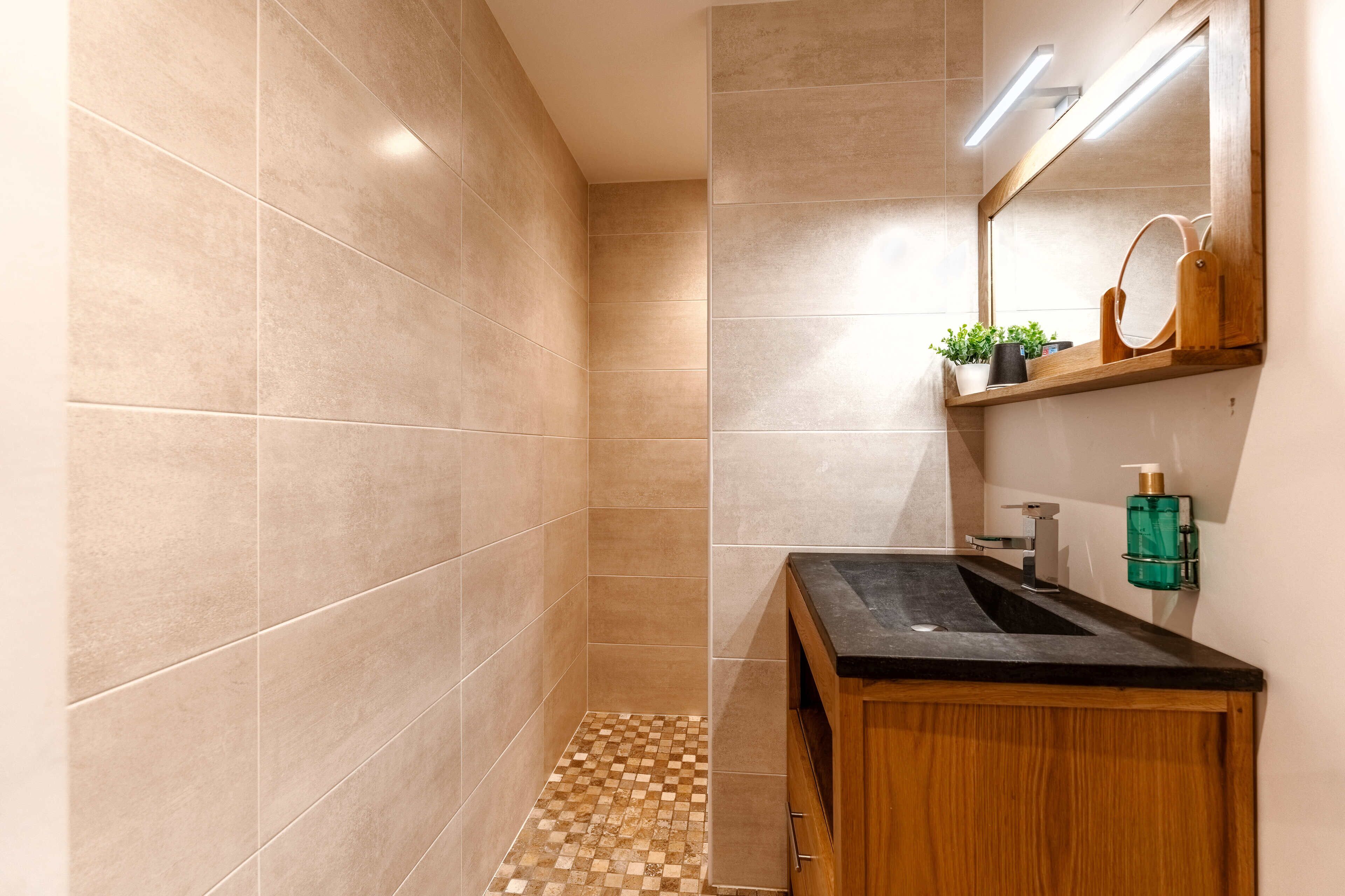 A modern bathroom with beige tiles, a wooden vanity, a sink, a mirror, and a small shelf with plants and toiletries.