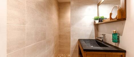 A modern bathroom with beige tiles, a wooden vanity, a sink, a mirror, and a small shelf with plants and toiletries.
