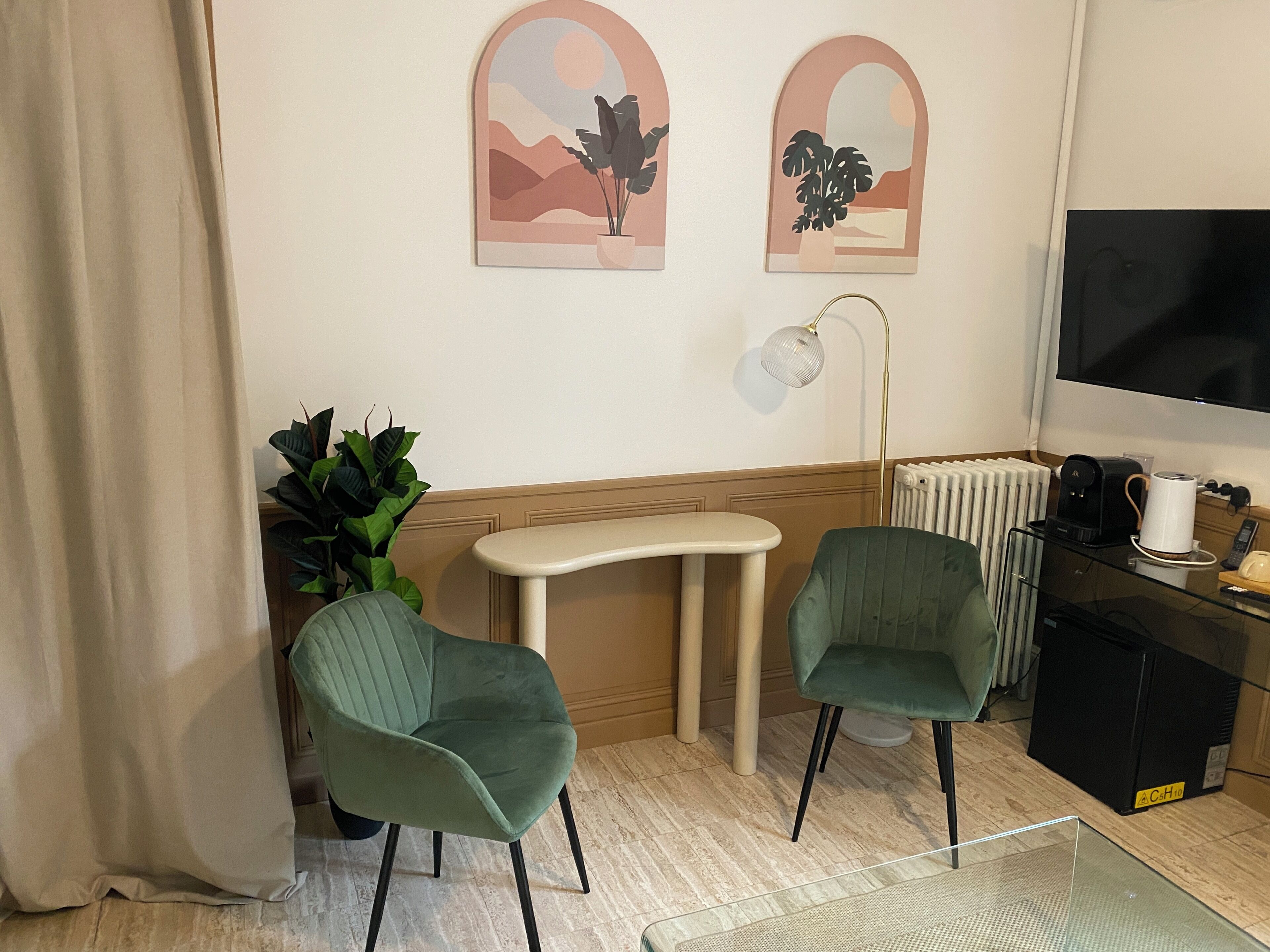 A room with two green chairs, a small table, a radiator, a coffee machine, and two wall-mounted artworks.