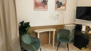 A room with two green chairs, a small table, a radiator, a coffee machine, and two wall-mounted artworks.
