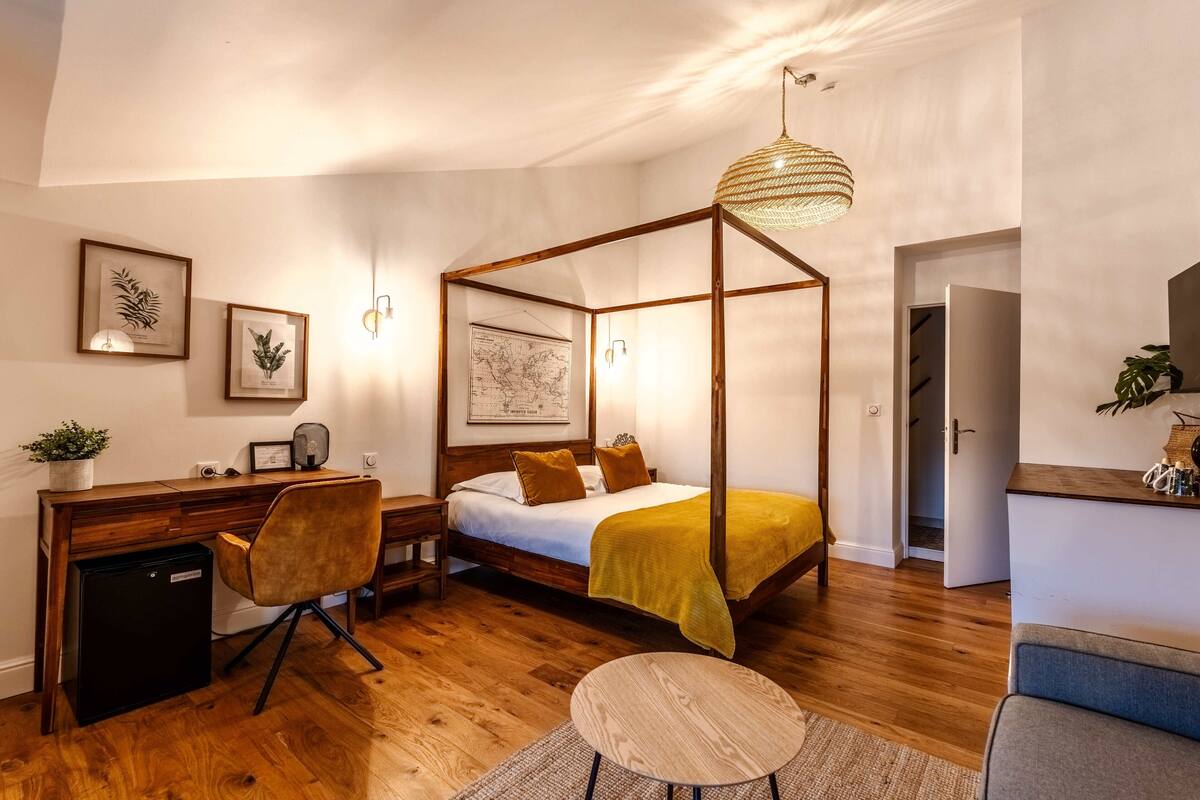 A bedroom with a four-poster bed, a desk with a chair, a small refrigerator, and a round table.