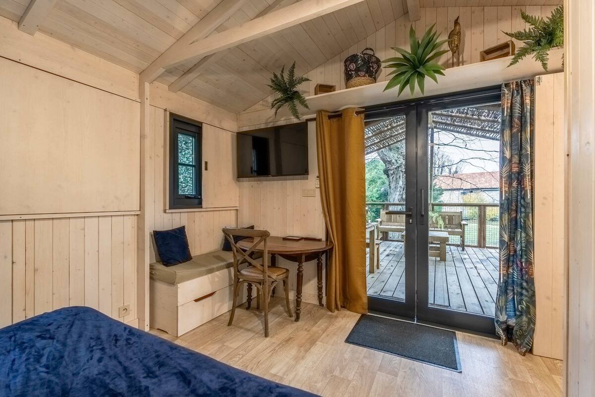 A cozy room with a bed, a desk, and a chair. There is a window, a door leading to an outdoor area, and a shelf with plants.
