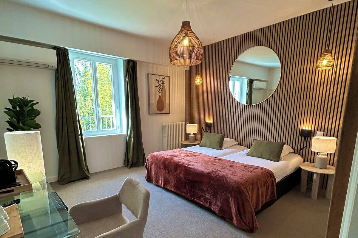 A hotel room with a bed, bedside lamps, a round mirror, a desk with a chair, and a window with curtains.