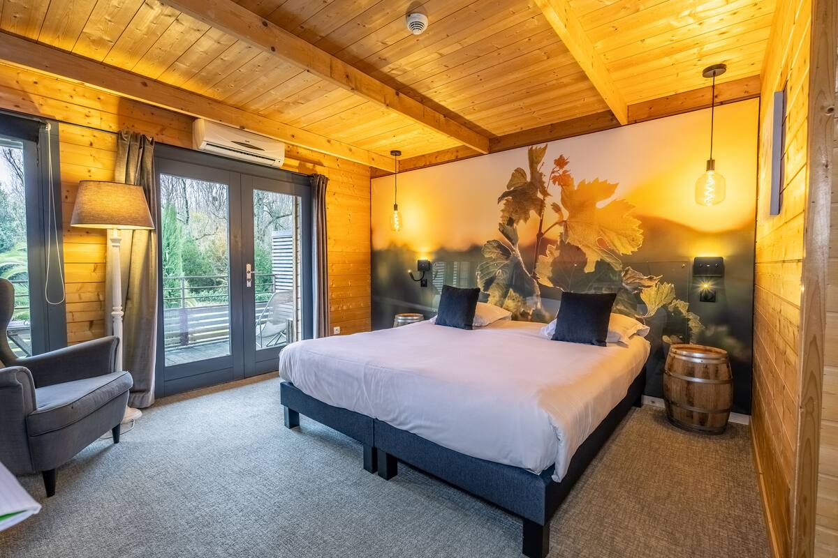 A wooden cabin bedroom with a large bed, a nightstand, a lamp, and a view through a sliding glass door.