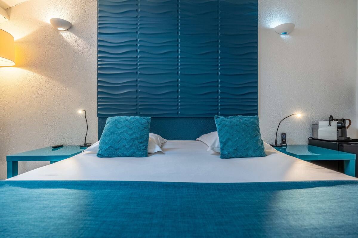 A hotel room with a large bed, two matching blue pillows, a nightstand with a lamp, and a wall-mounted light fixture.