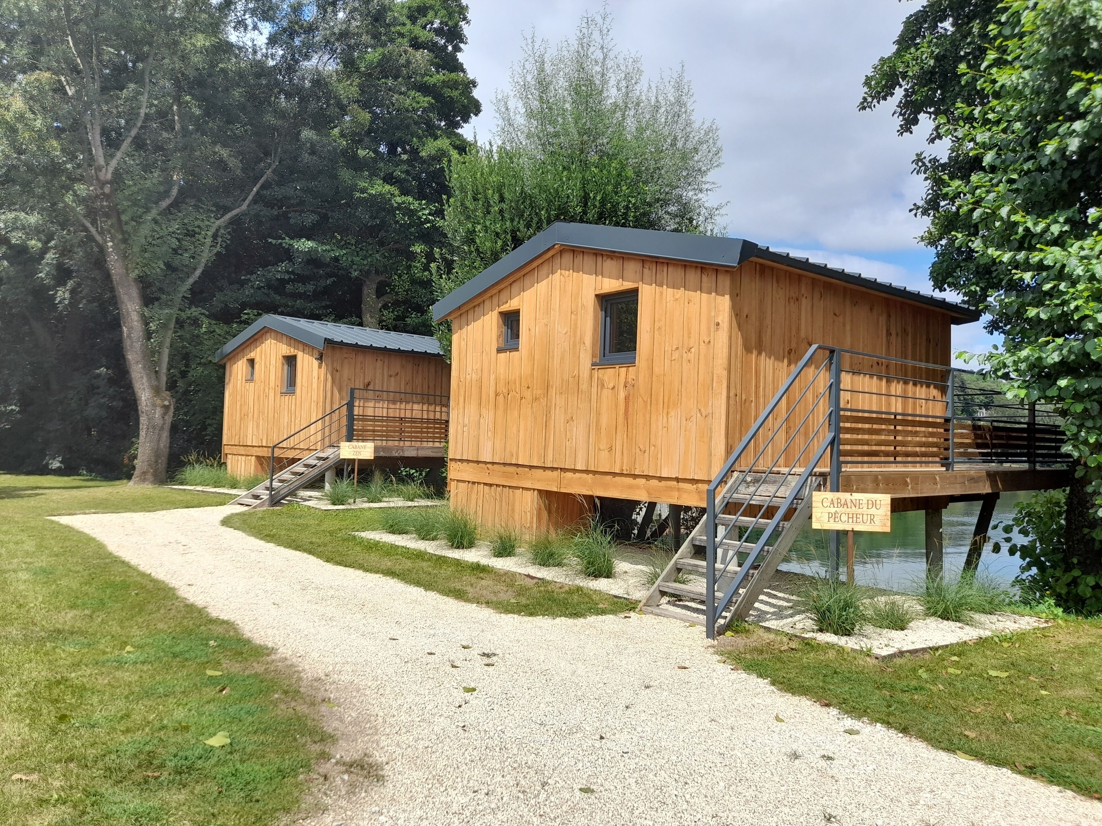 Two wooden cabins with external staircases, a gravel path, and a sign indicating 'Cabane du pêcheur'.