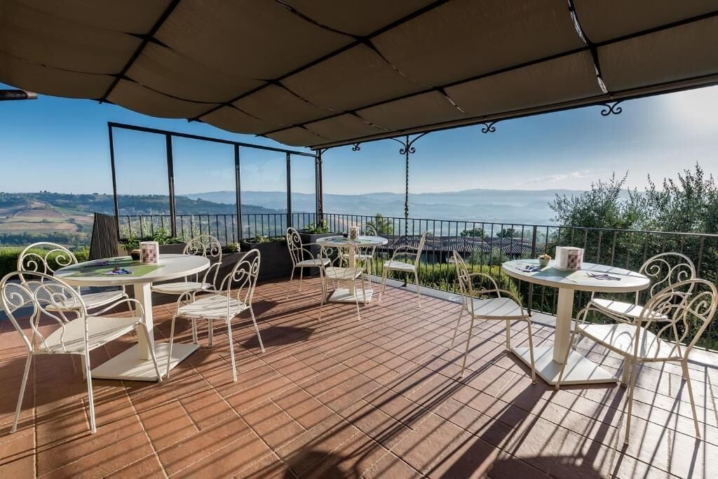 Un patio al aire libre con dos mesas redondas y sillas, una pérgola en la parte superior y una vista panorámica de colinas y montañas.