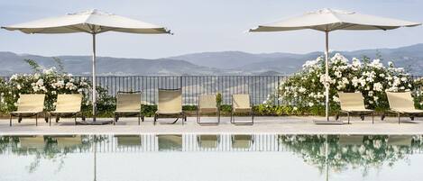 A poolside area with lounge chairs and umbrellas, a clear view of the mountains, and a calm water surface reflecting the surroundings.
