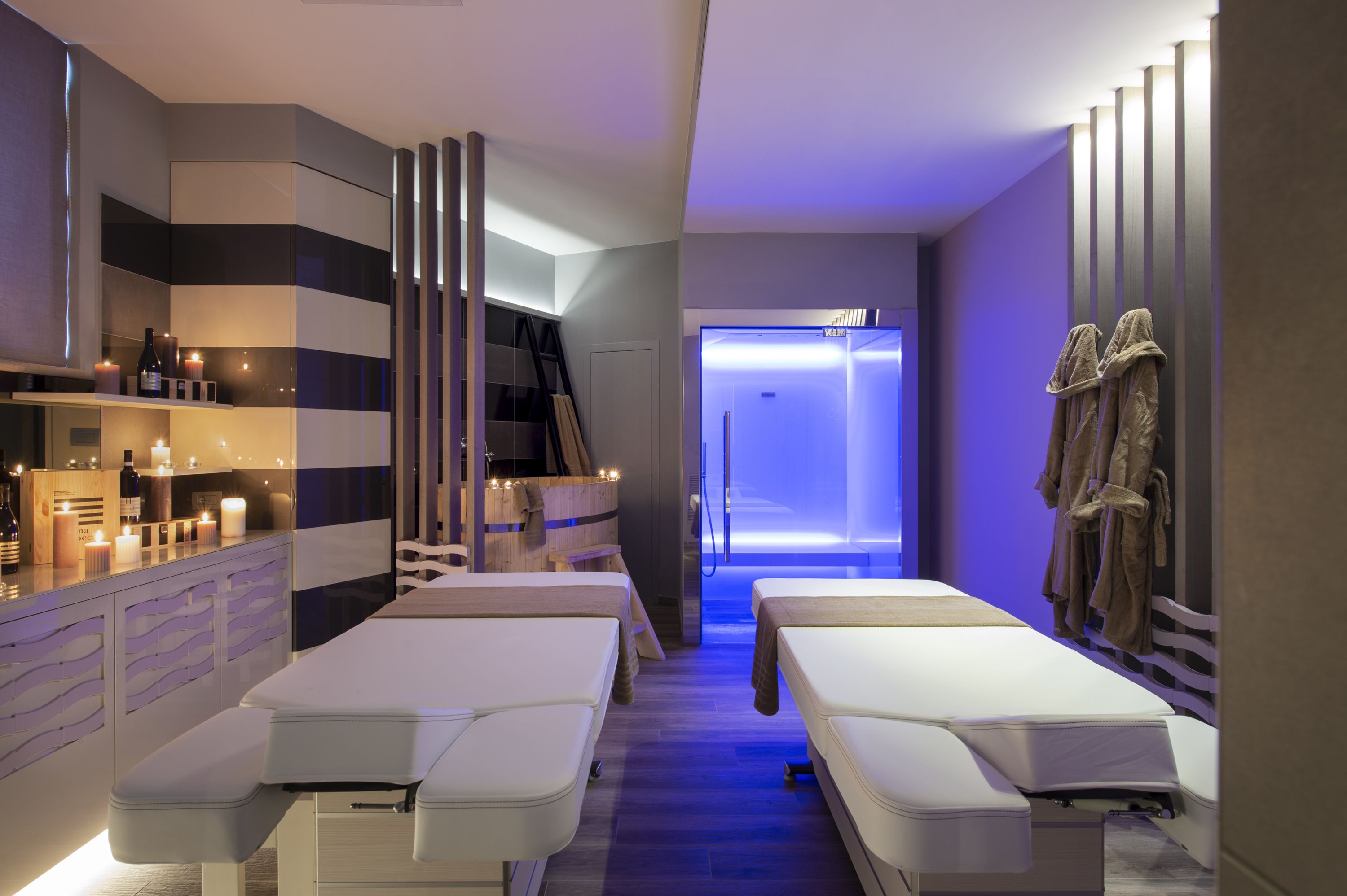 Un spa moderno con dos camillas de masaje, una zona de recepción con velas y una entrada iluminada en azul.