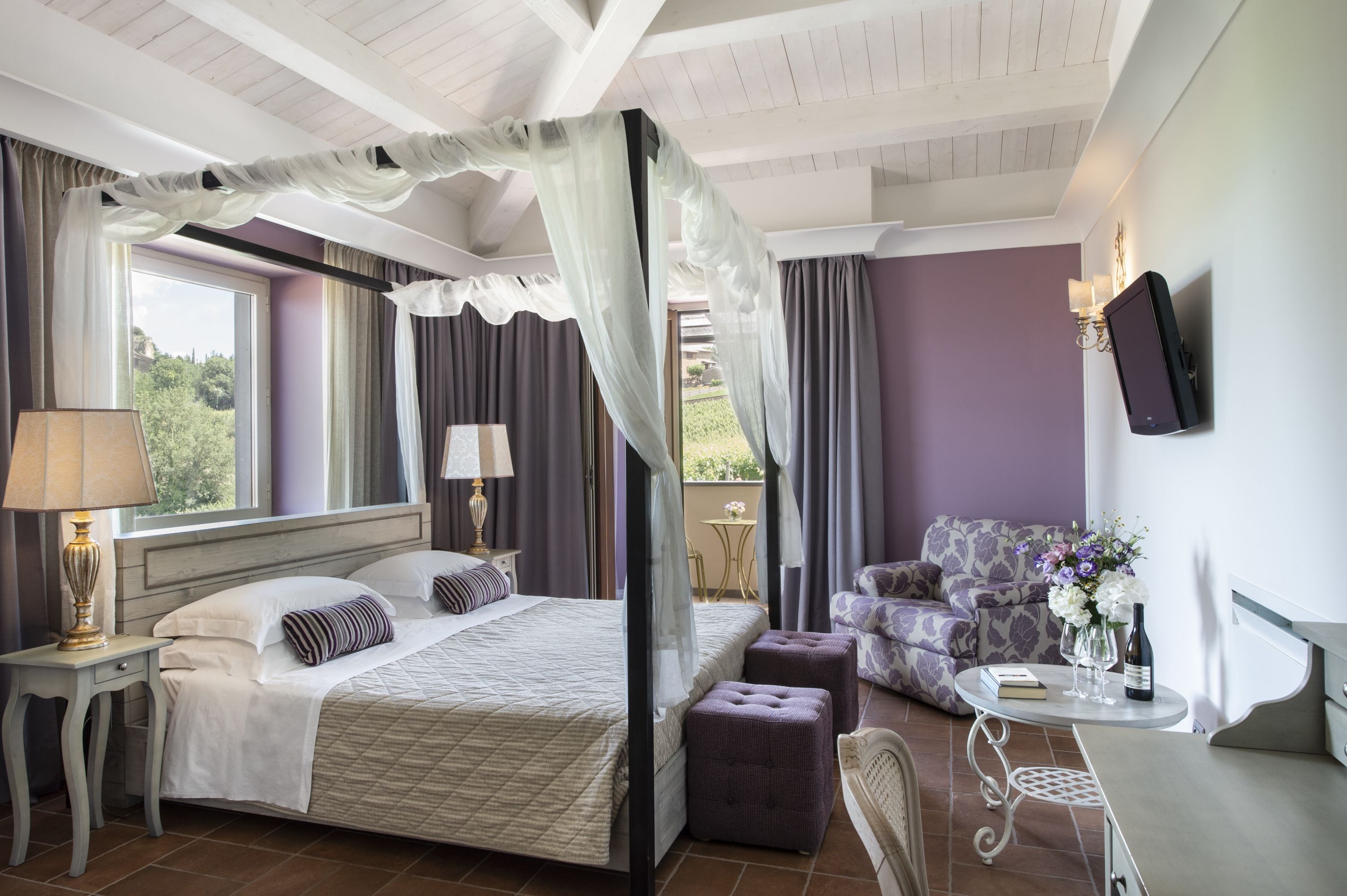 Une chambre à coucher avec un lit à baldaquin, un coin salon avec un canapé, une petite table sur laquelle sont posés un vase et une bouteille de vin, et un téléviseur fixé au mur.