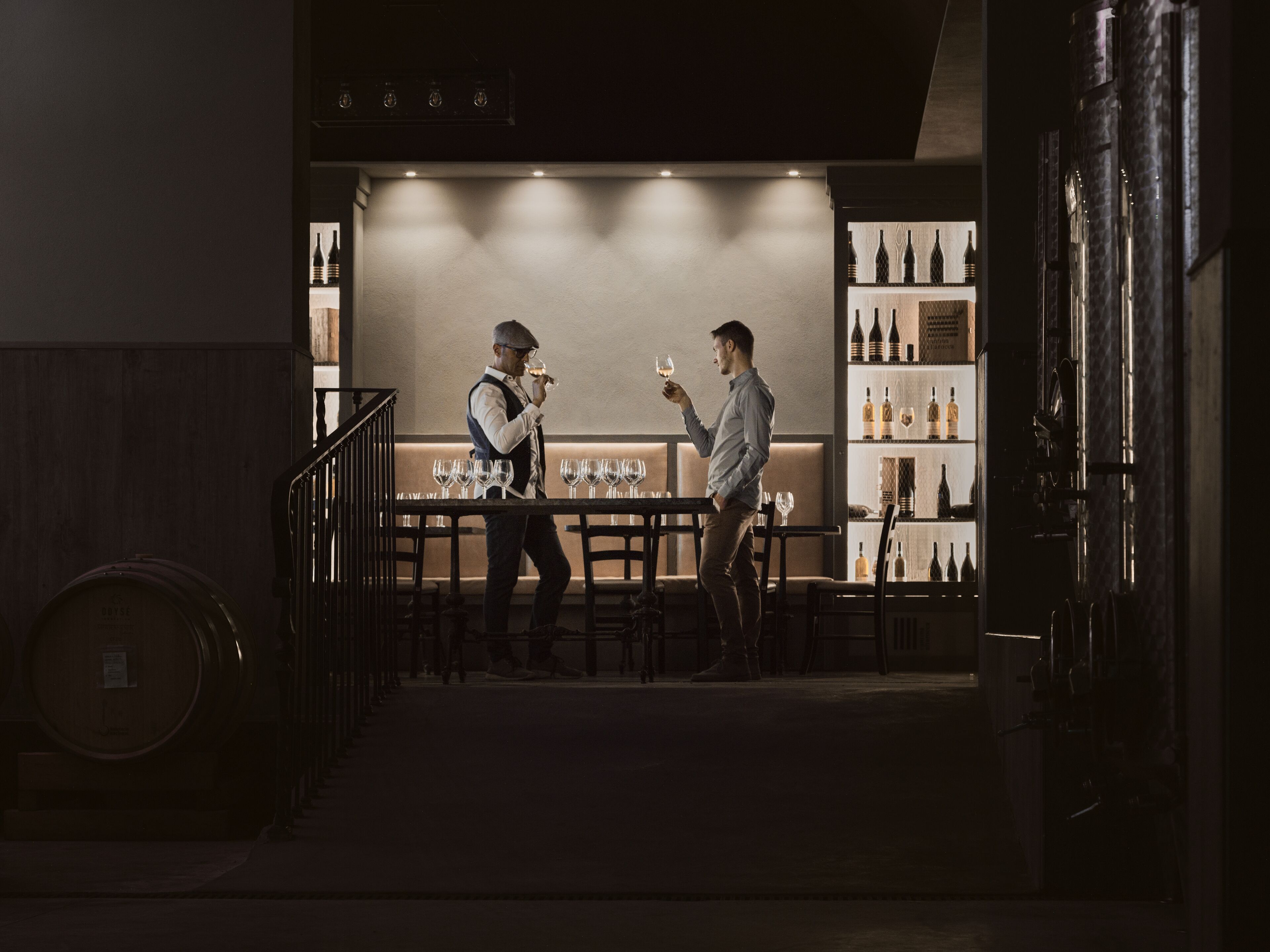 Un coin bar à la lumière tamisée où deux hommes sont en conversation, avec un présentoir à bouteilles de vin et un escalier menant à un niveau inférieur.
