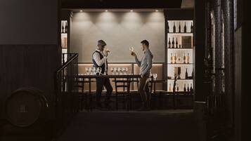 Un coin bar à la lumière tamisée où deux hommes sont en conversation, avec un présentoir à bouteilles de vin et un escalier menant à un niveau inférieur.
