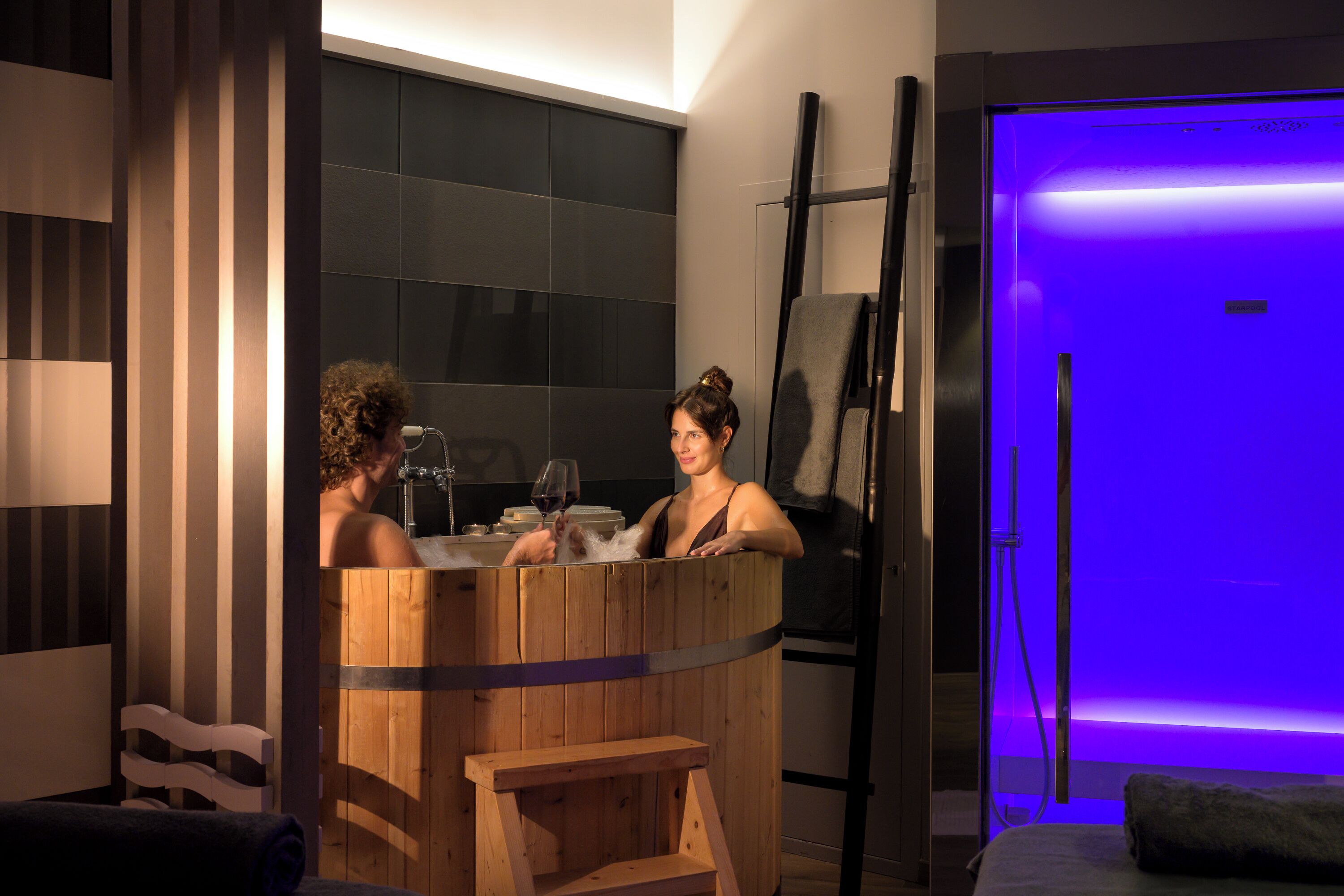 Eine hölzerne Badewanne mit einem Weinglas, einem Handtuch und einer Sitzbank in einem modernen Badezimmer mit dunklen Fliesen und einer blauen Wandbeleuchtung.