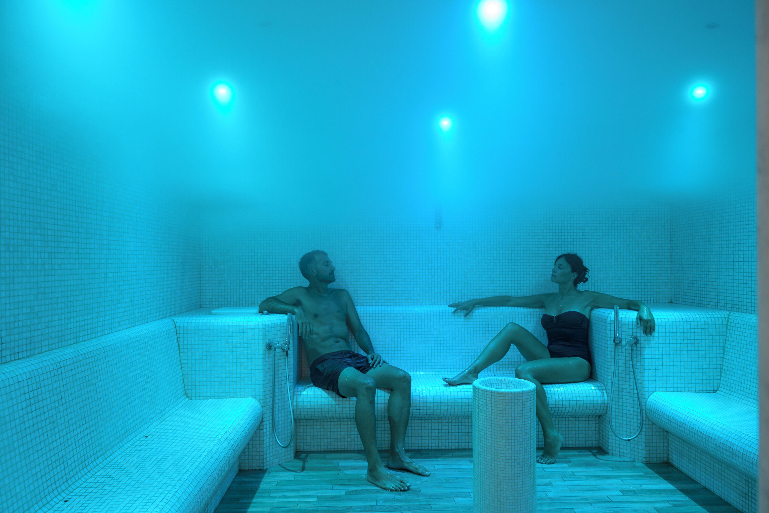 A room with two white cushioned seating areas, a tiled floor, and a ceiling with recessed lighting.