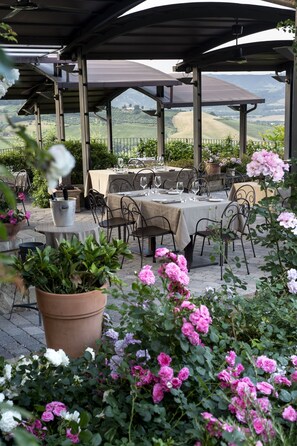 Un área de comedor al aire libre con mesas y sillas, rodeada de plantas y flores en macetas.
