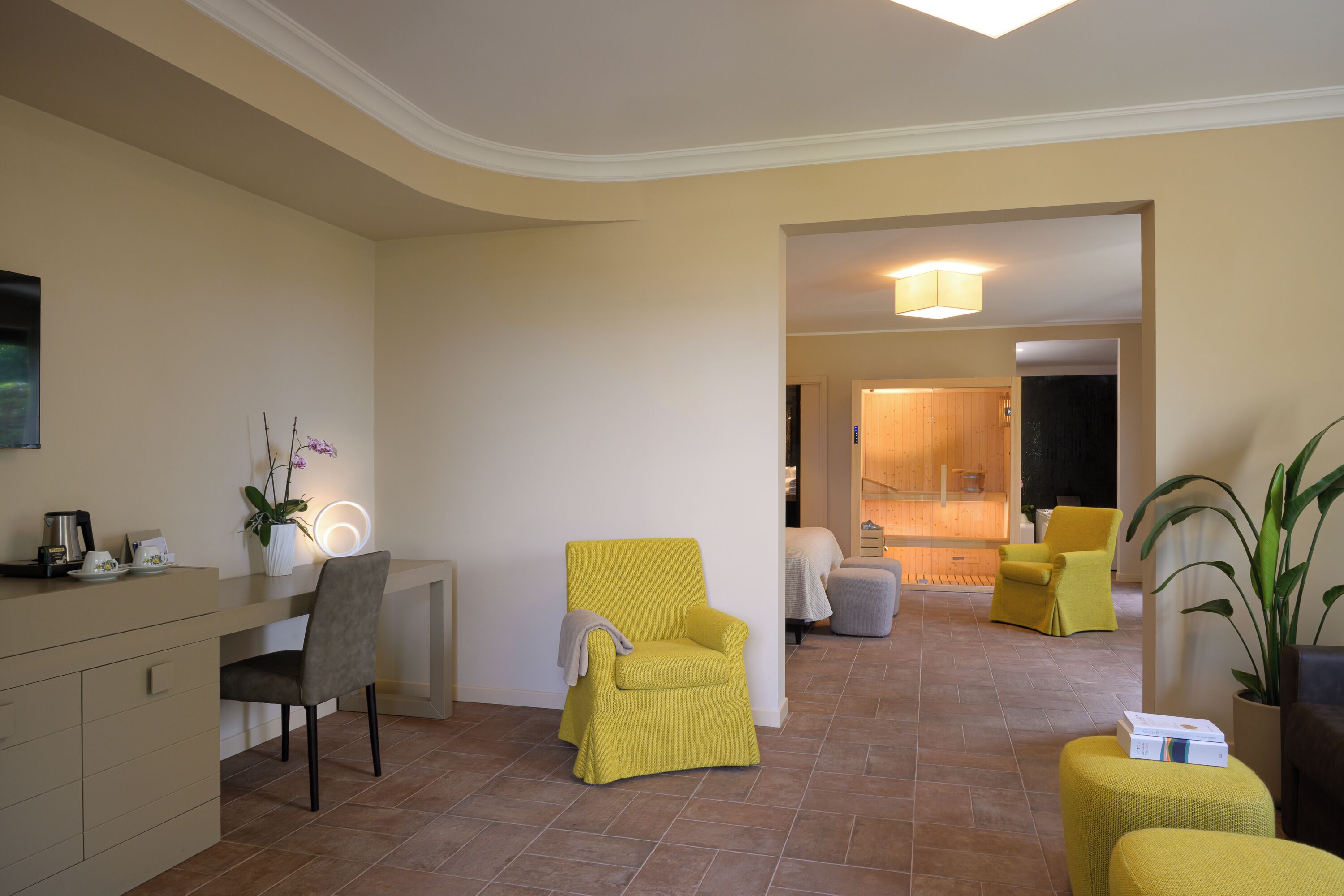 Un salon moderne avec un fauteuil jaune, un canapé gris et une petite table avec une lampe. On aperçoit à l’arrière-plan un coin cuisine avec un meuble en bois et un plan de travail.