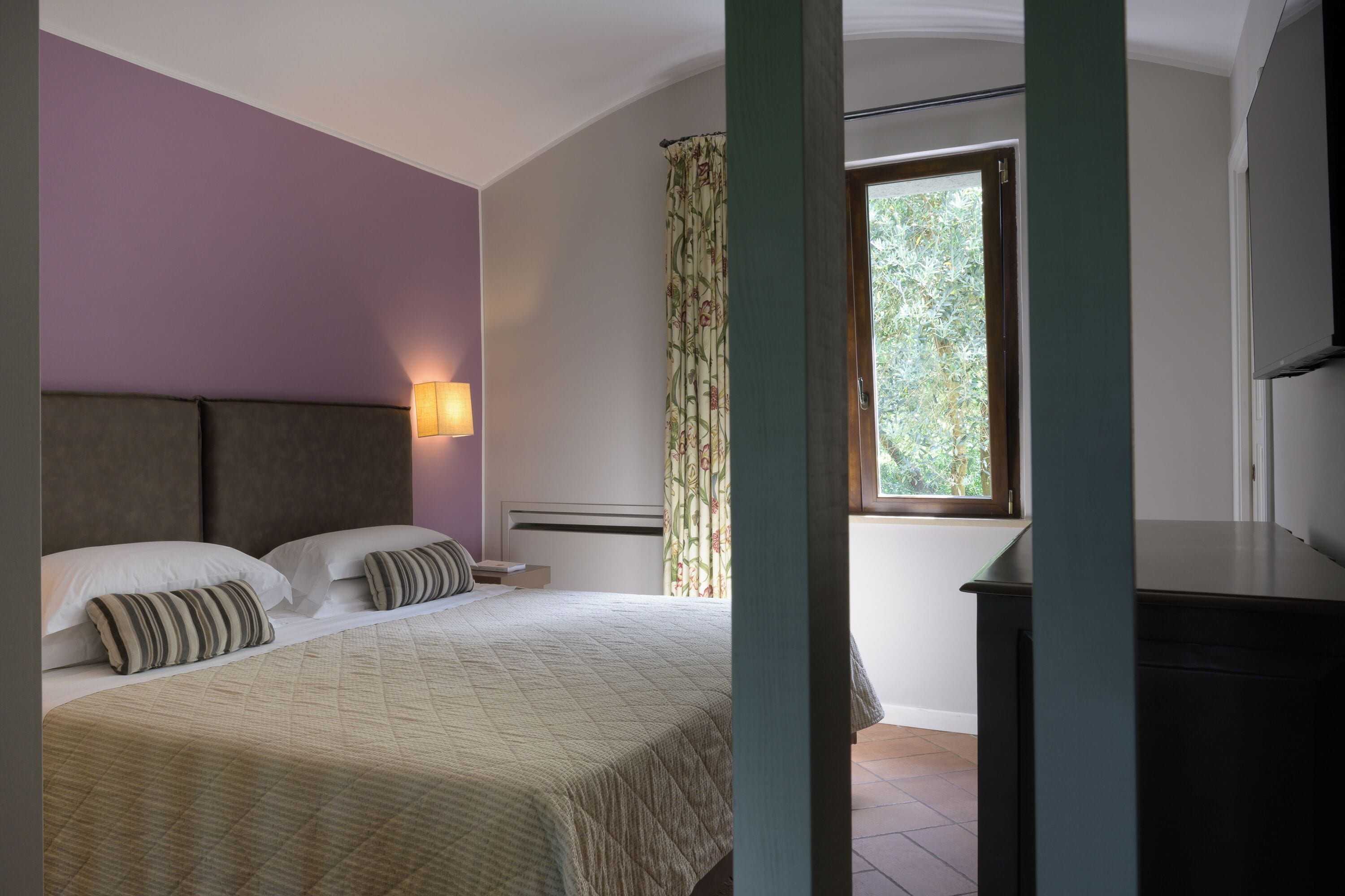 A bedroom with a bed, a nightstand, a lamp, a window with curtains, and a view of greenery outside.