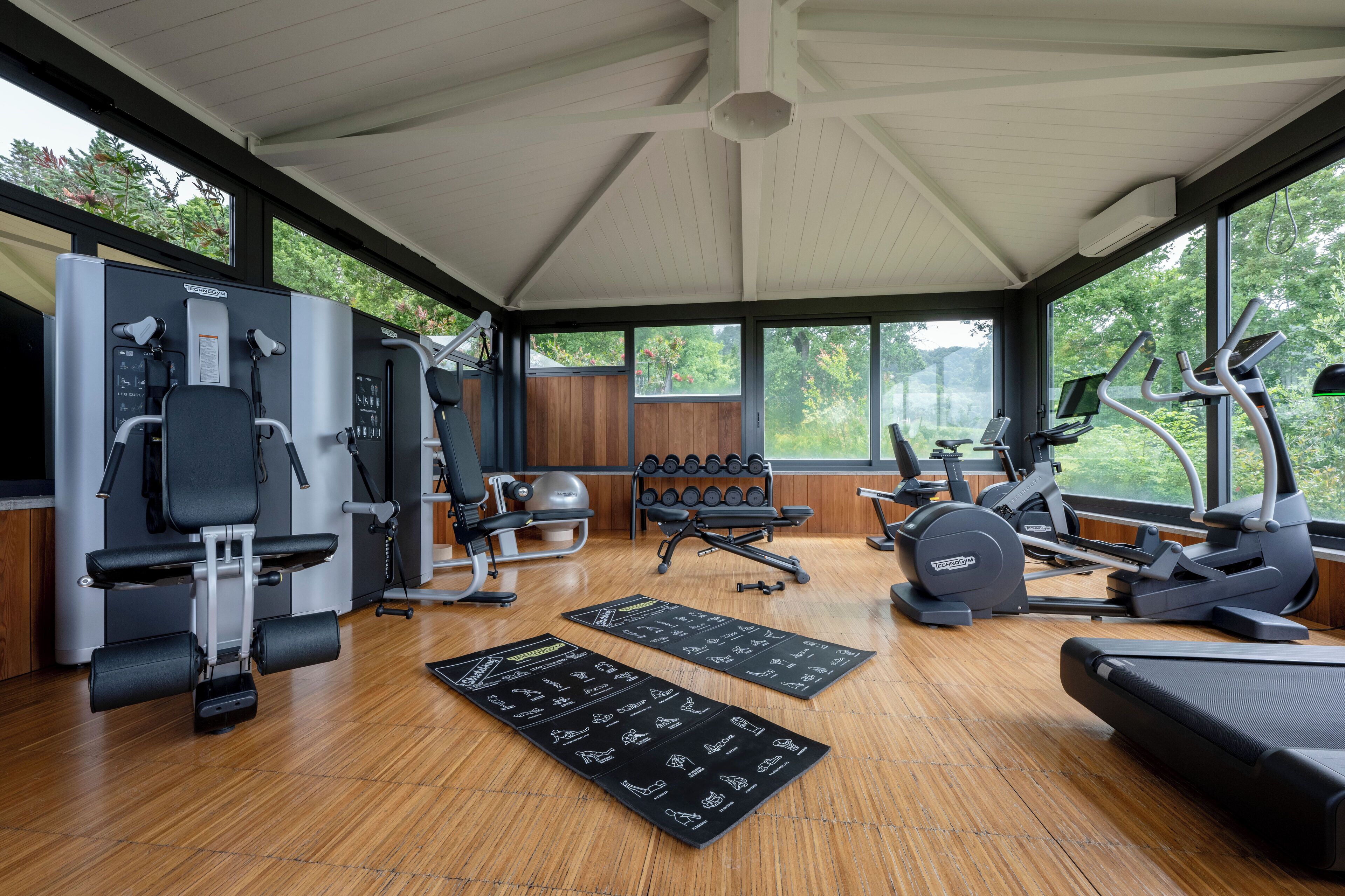 A gym with various exercise equipment, a bench, and a large window offering a view of trees.