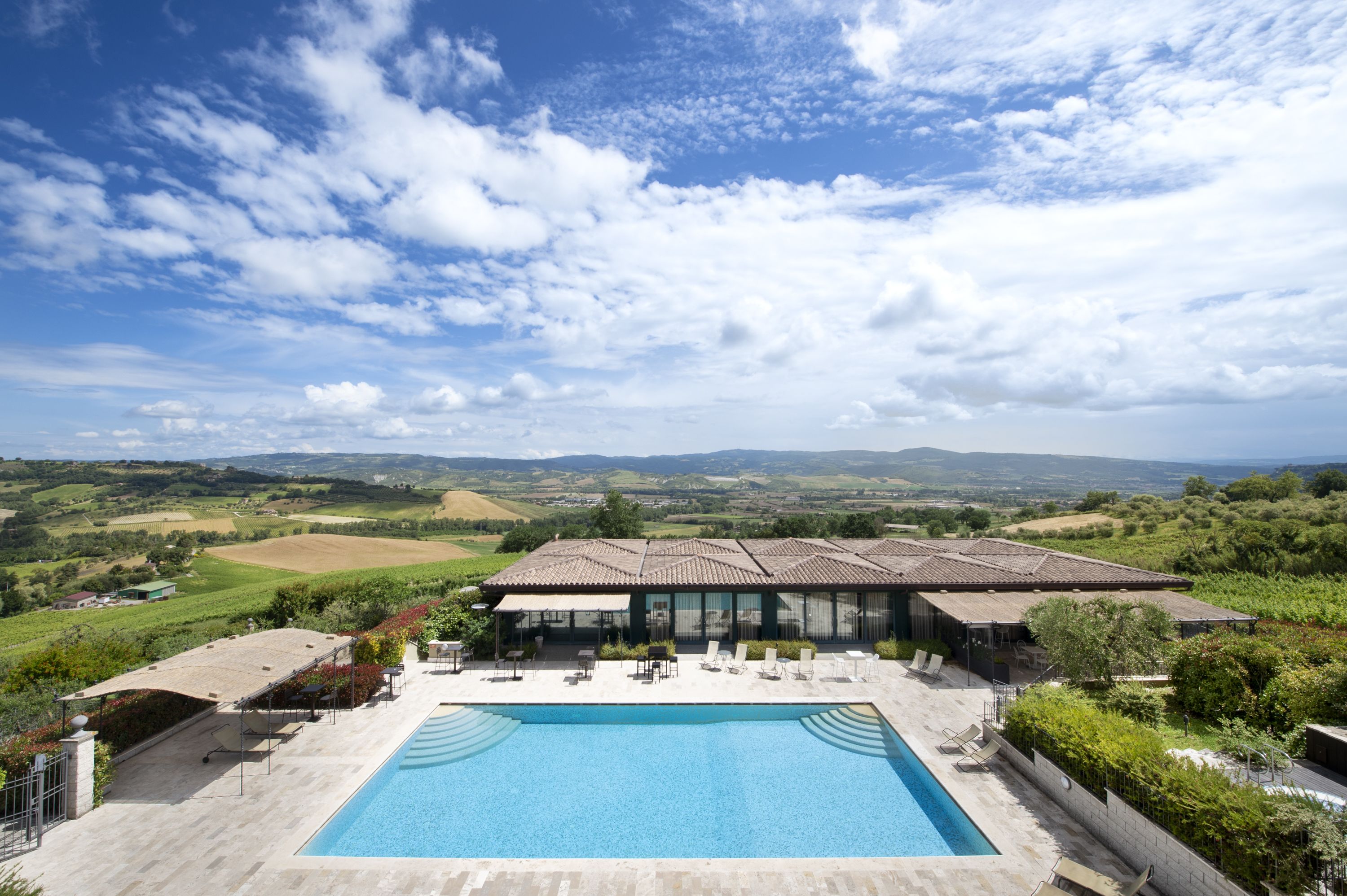 A large swimming pool with a patio area and lounge chairs, surrounded by lush greenery and a view of rolling hills.