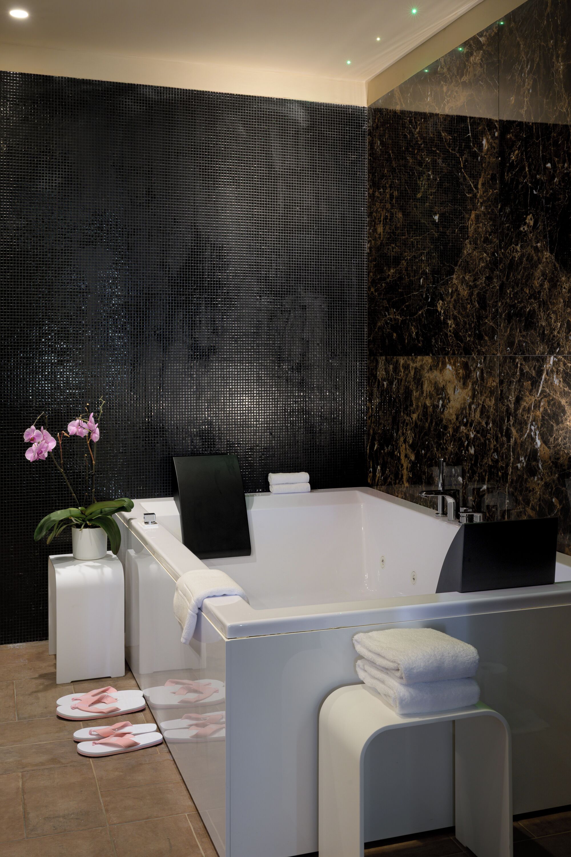 Un baño moderno con una bañera grande, un lavabo blanco y un espejo. Hay una orquídea en maceta y un par de pantuflas rosas en el suelo.
