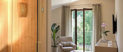 A modern room with a wooden sauna, a comfortable chair, a potted plant, and a large window offering a view of greenery.