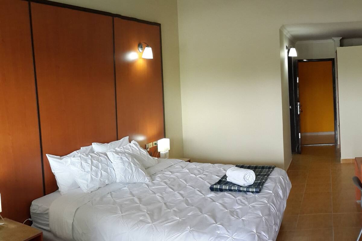 A hotel room with a large bed, a wooden headboard, two bedside lamps, and a bathroom visible in the background.