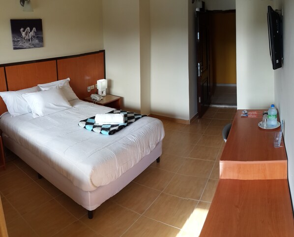 A hotel room with a bed, a nightstand with a phone, a checkered blanket, and a desk with a water bottle and glasses.