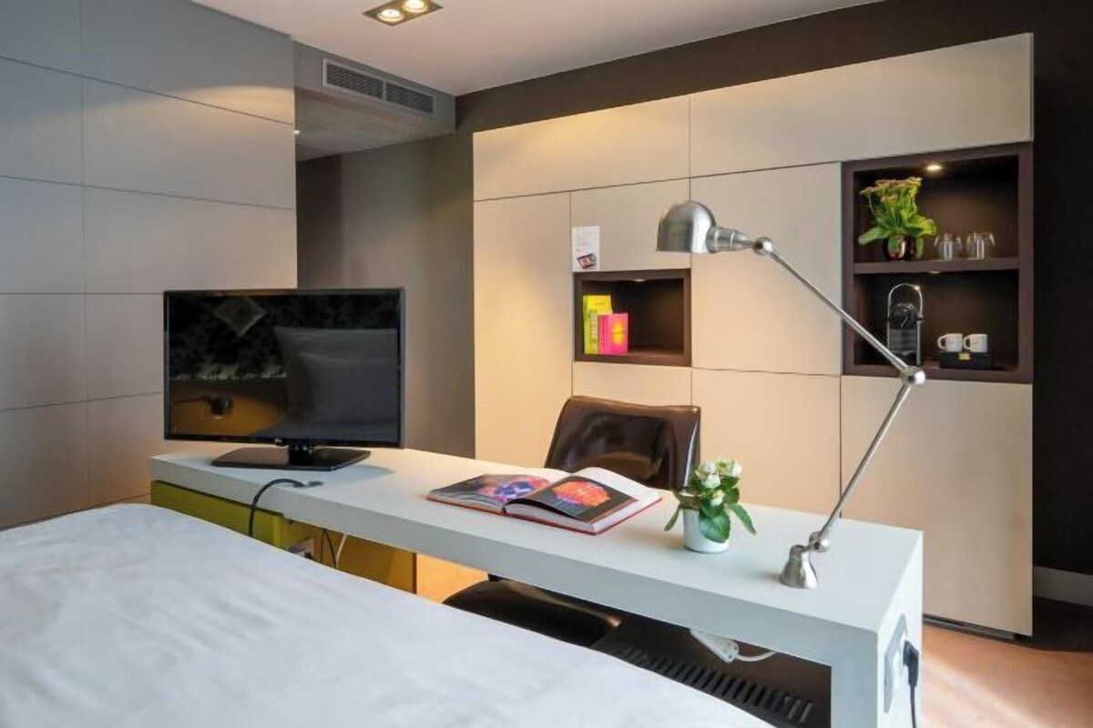 A modern hotel room with a bed, a desk with a computer, a TV, a lamp, and a shelf with decorative items.