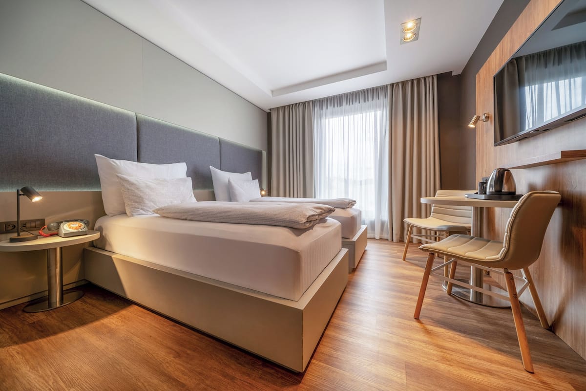 A modern hotel room with a large bed, bedside table, lamp, and a desk with a chair.
