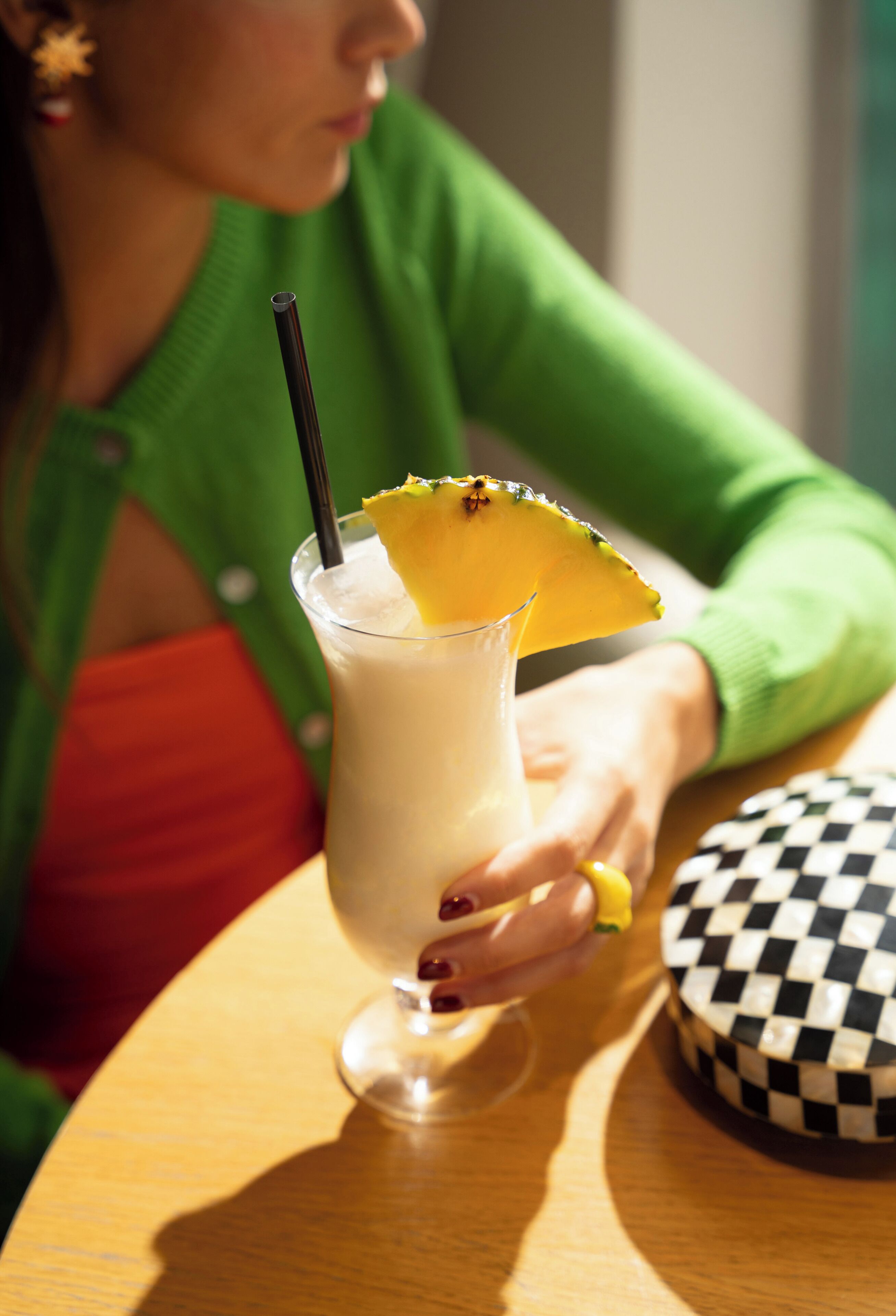Una persona sosteniendo un vaso con pajita y una rodaja de fruta, sentada a una mesa con un objeto a cuadros al lado.