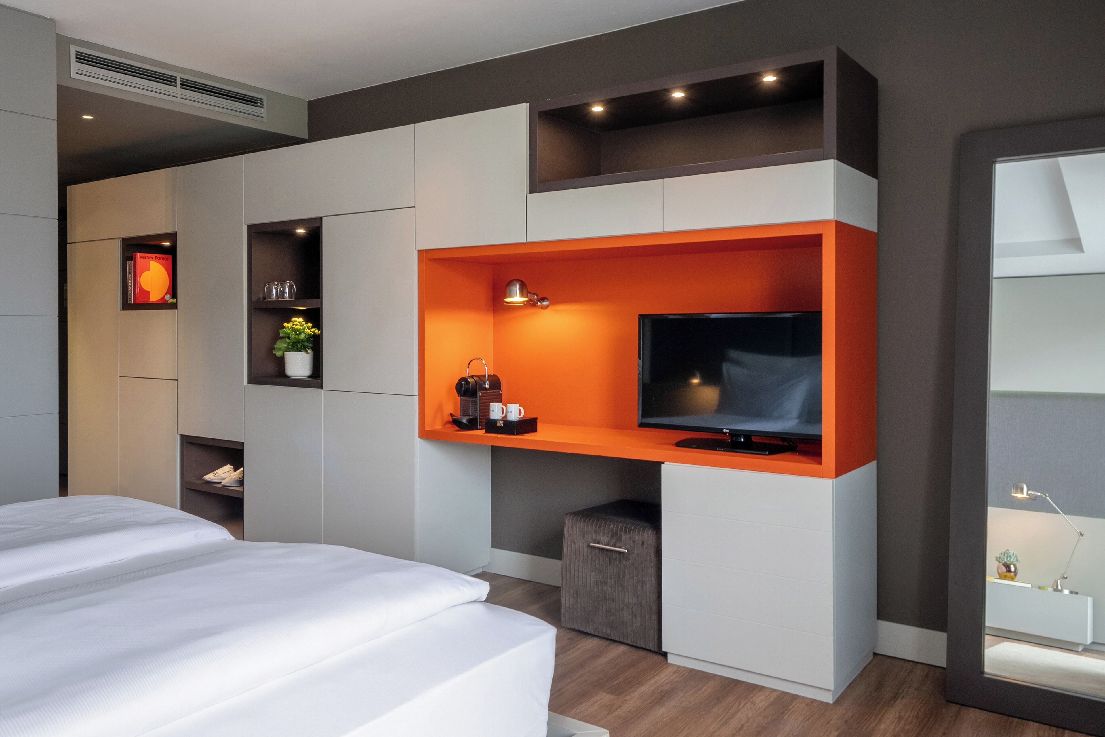 Una habitación de hotel moderna con una cama, un televisor instalado en una pared anaranjada de acento y una pequeña cafetera, además de una planta.