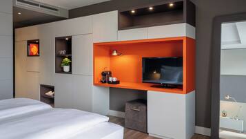Una habitación de hotel moderna con una cama, un televisor instalado en una pared anaranjada de acento y una pequeña cafetera, además de una planta.