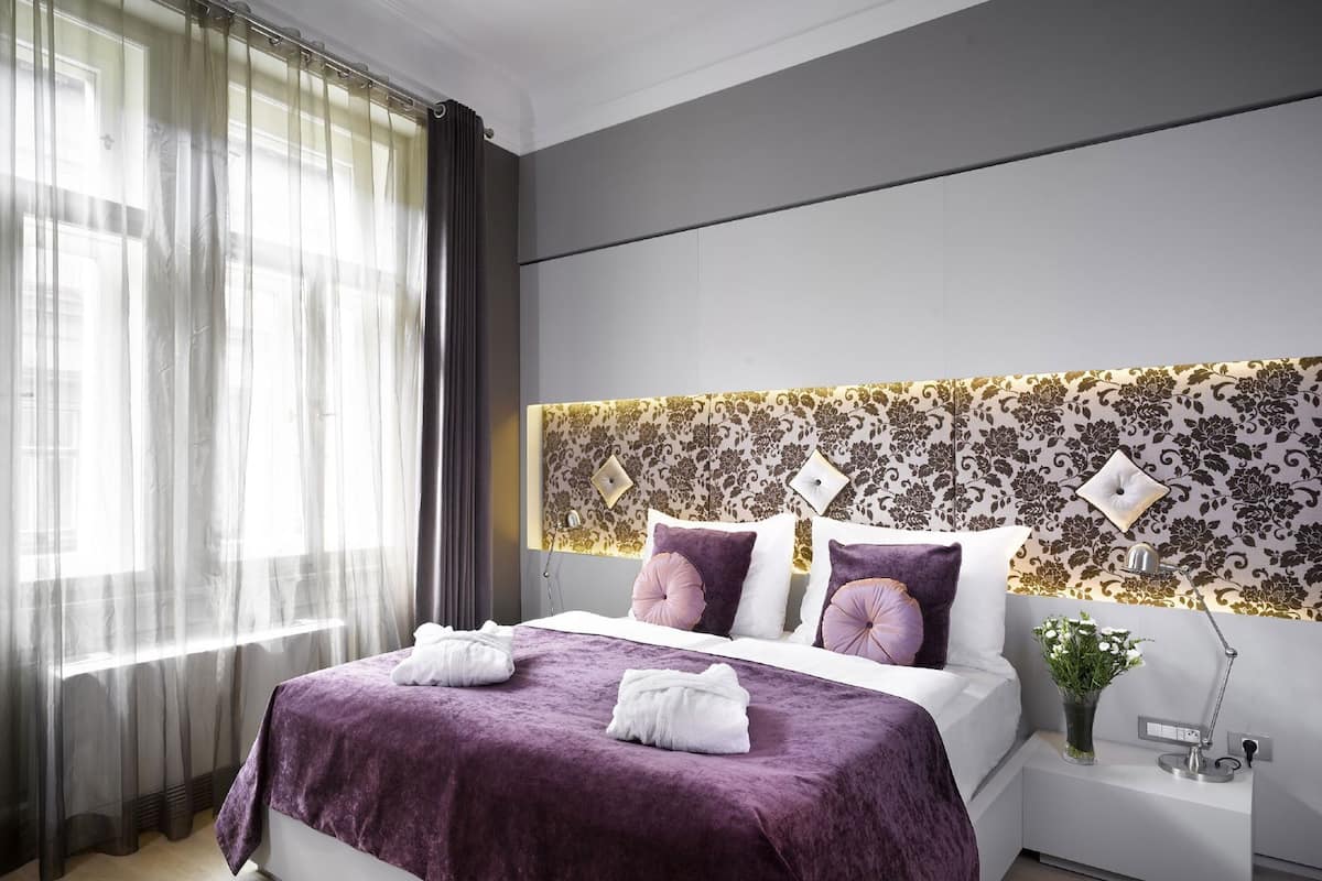 A modern hotel room with a large bed, a headboard with a floral pattern, a bedside table with a lamp, and sheer curtains on the window.