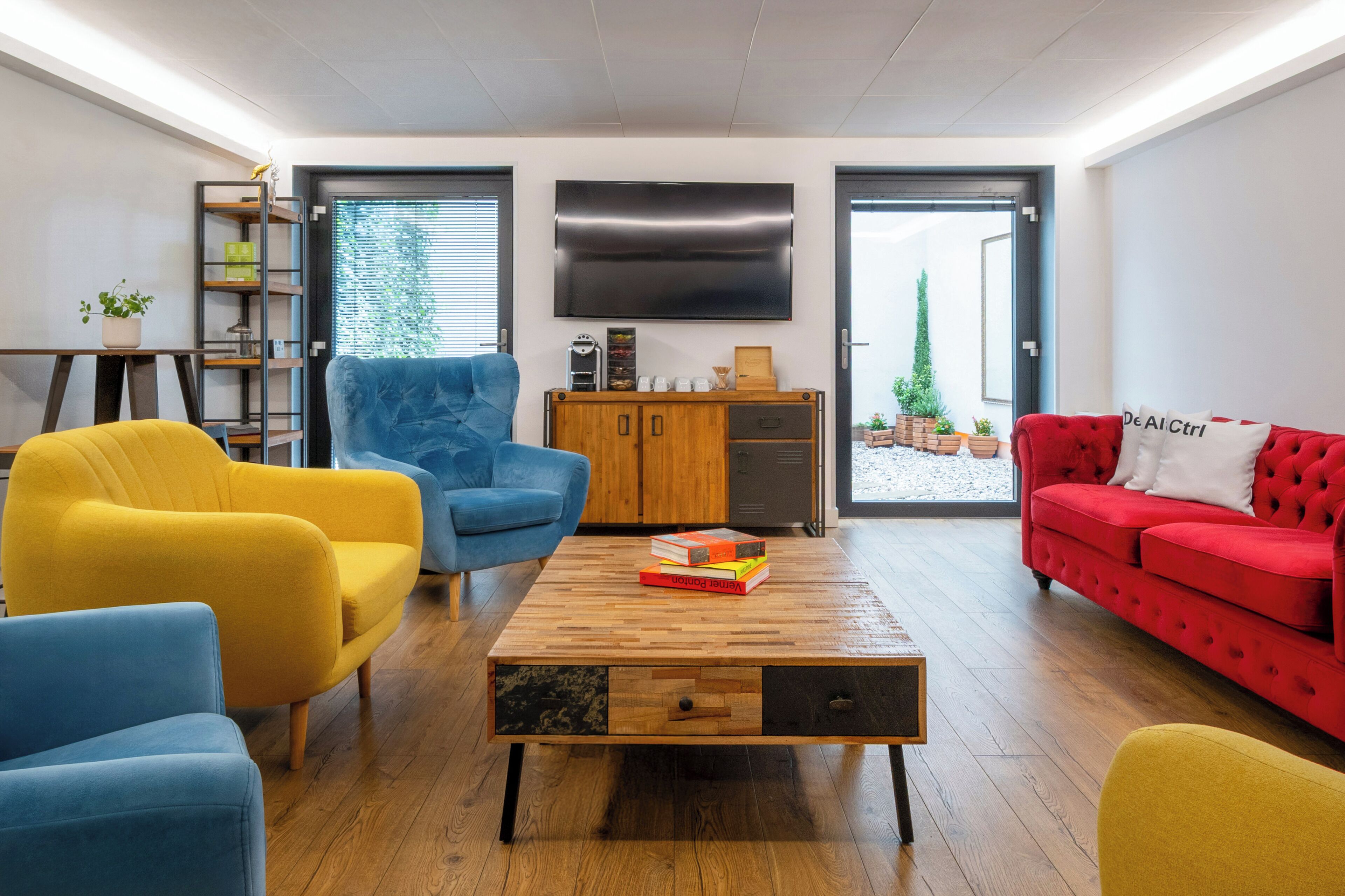 Una sala de estar moderna con una mesa de centro de madera, sillones azul y amarillo, un sofá rojo y un televisor montado en la pared.