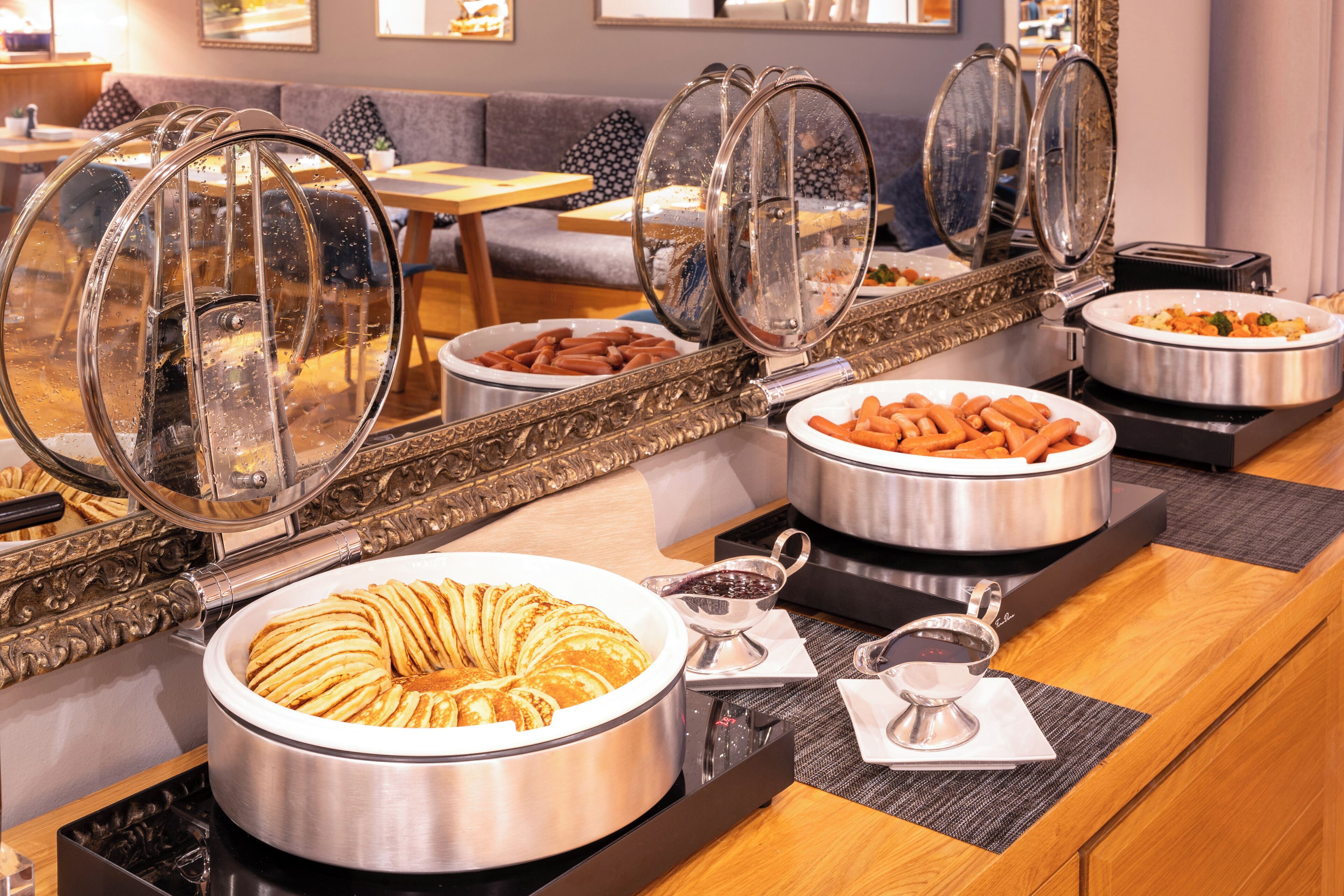A buffet setup with various food items, including a large pot of pancakes, a bowl of sausages, and a container of hot dogs.