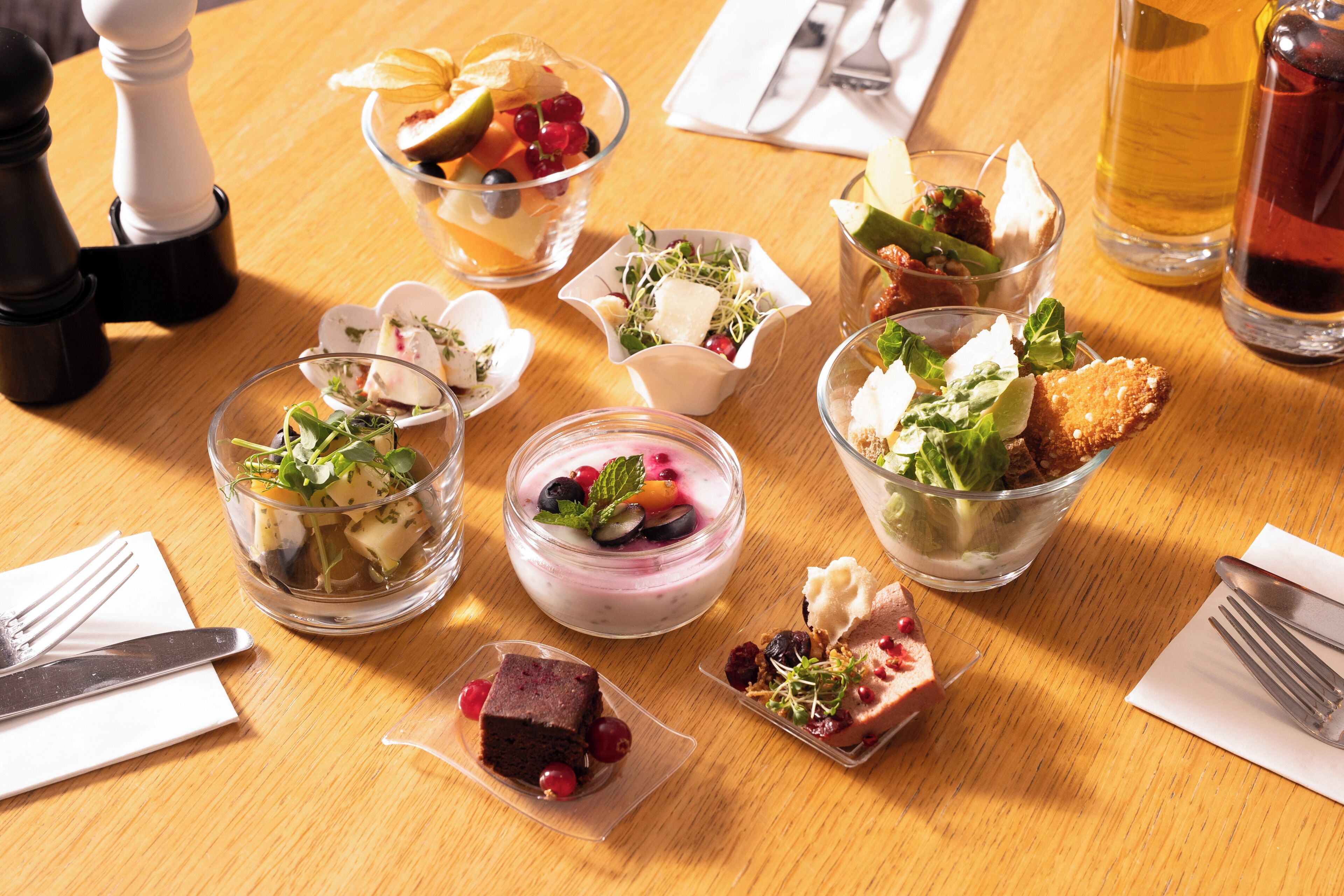 Diversos pratos e bebidas sobre uma mesa de madeira, incluindo uma salada em uma tigela, um copo de suco e um copo de cerveja.