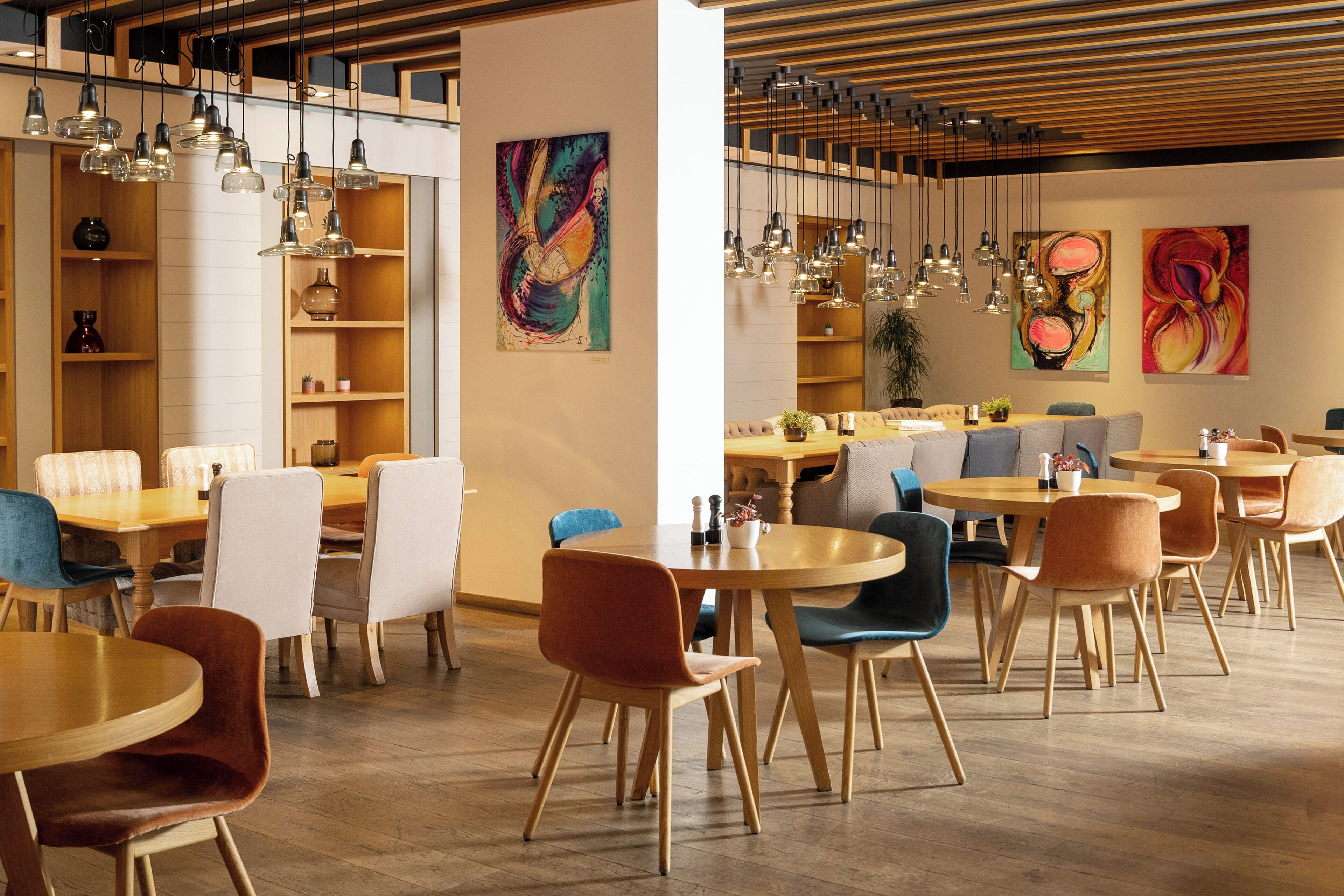 Een moderne eetruimte met houten tafels en stoelen, hanglampen en abstracte kunst aan de muren.