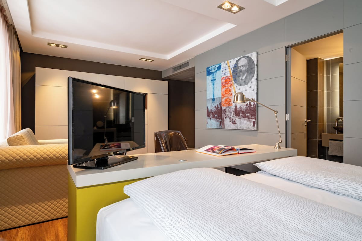 A modern hotel room with a bed, a desk with a computer, a chair, and a large poster on the wall.