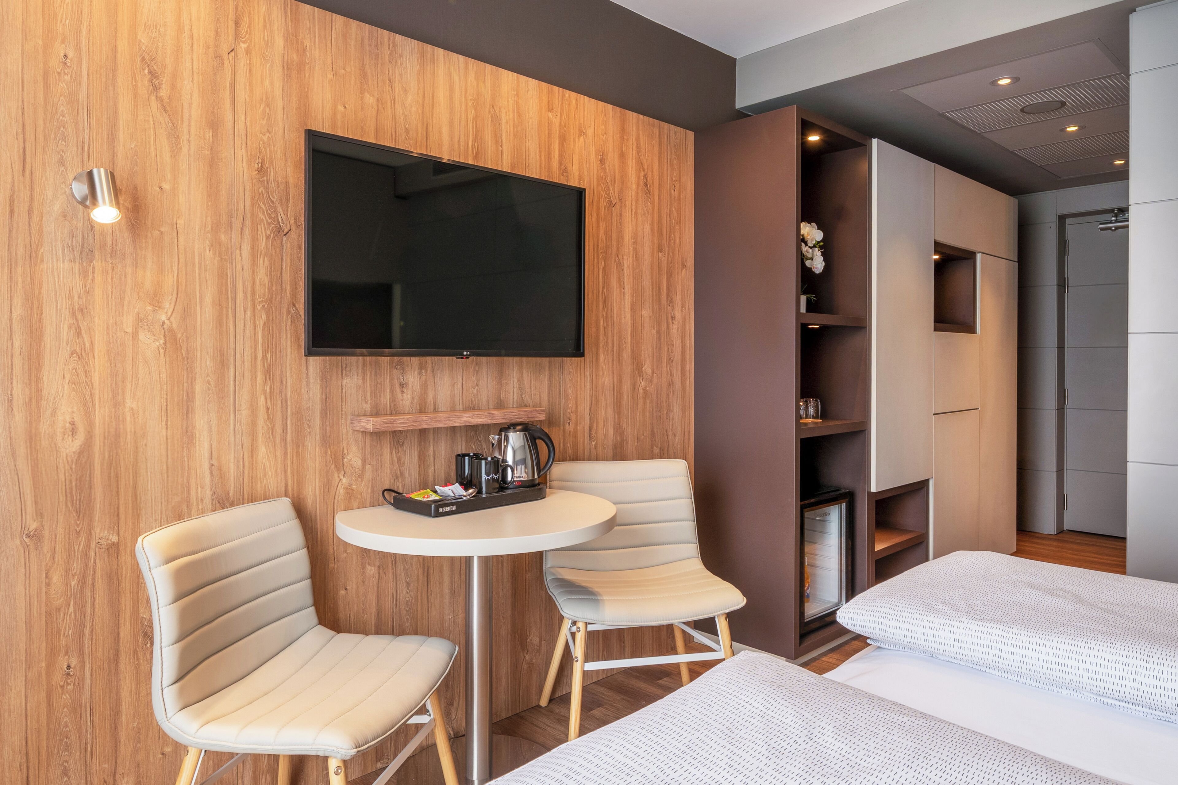 Hotellihuone, jossa on puinen seinä, litteänäyttöinen televisio, kaksi pehmustettua tuolia, pieni pyöreä pöytä, jossa on kahvinkeitin, sekä valkoisella patjalla varustettu sänky.