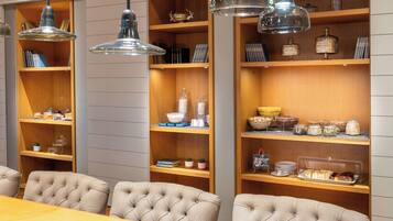 Un espace repas avec une table en bois et des chaises à capitonnage, entouré d’étagères où sont exposés des livres, des objets décoratifs et des produits alimentaires.