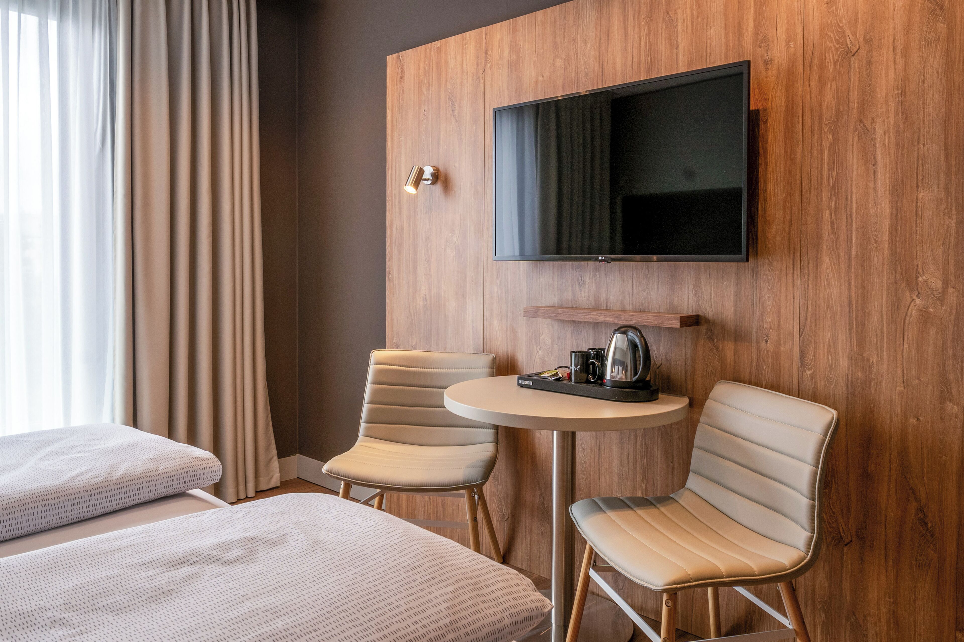 Hotelkamer met twee bedden, een aan de muur bevestigde televisie, een klein rond tafeltje met waterkoker en twee stoelen.
