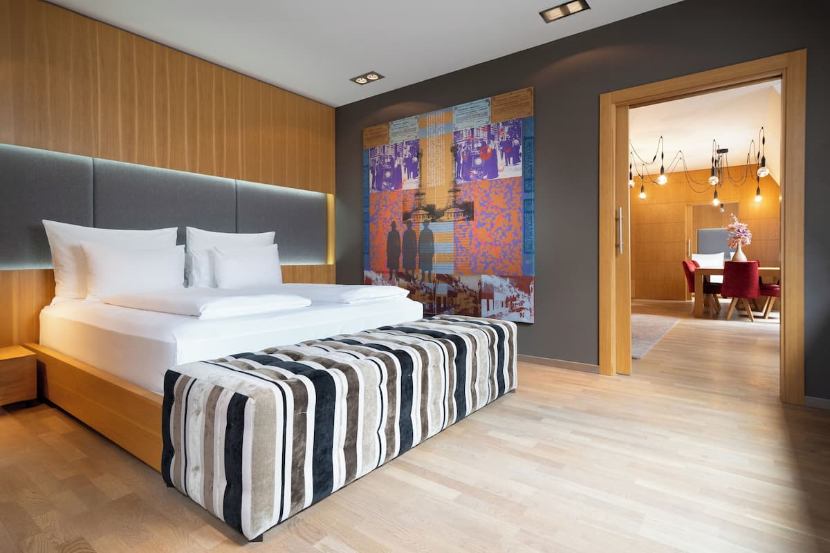 A modern hotel room with a large bed, a wooden headboard, a colorful artwork on the wall, and a dining area visible through an open door.