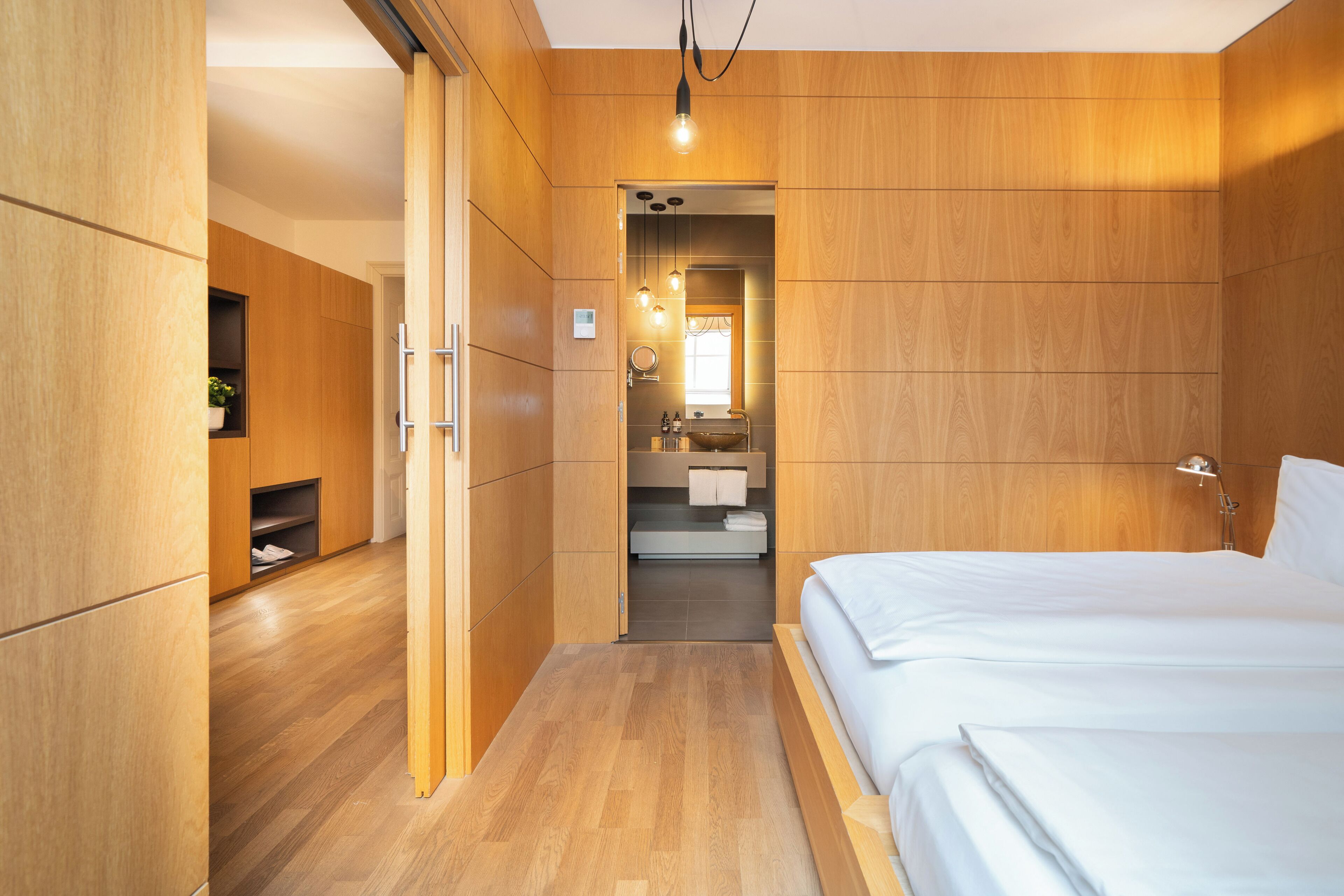 Una habitación de hotel moderna con paneles de madera, una cama grande, un armario empotrado y un baño con lavamanos y espejo.