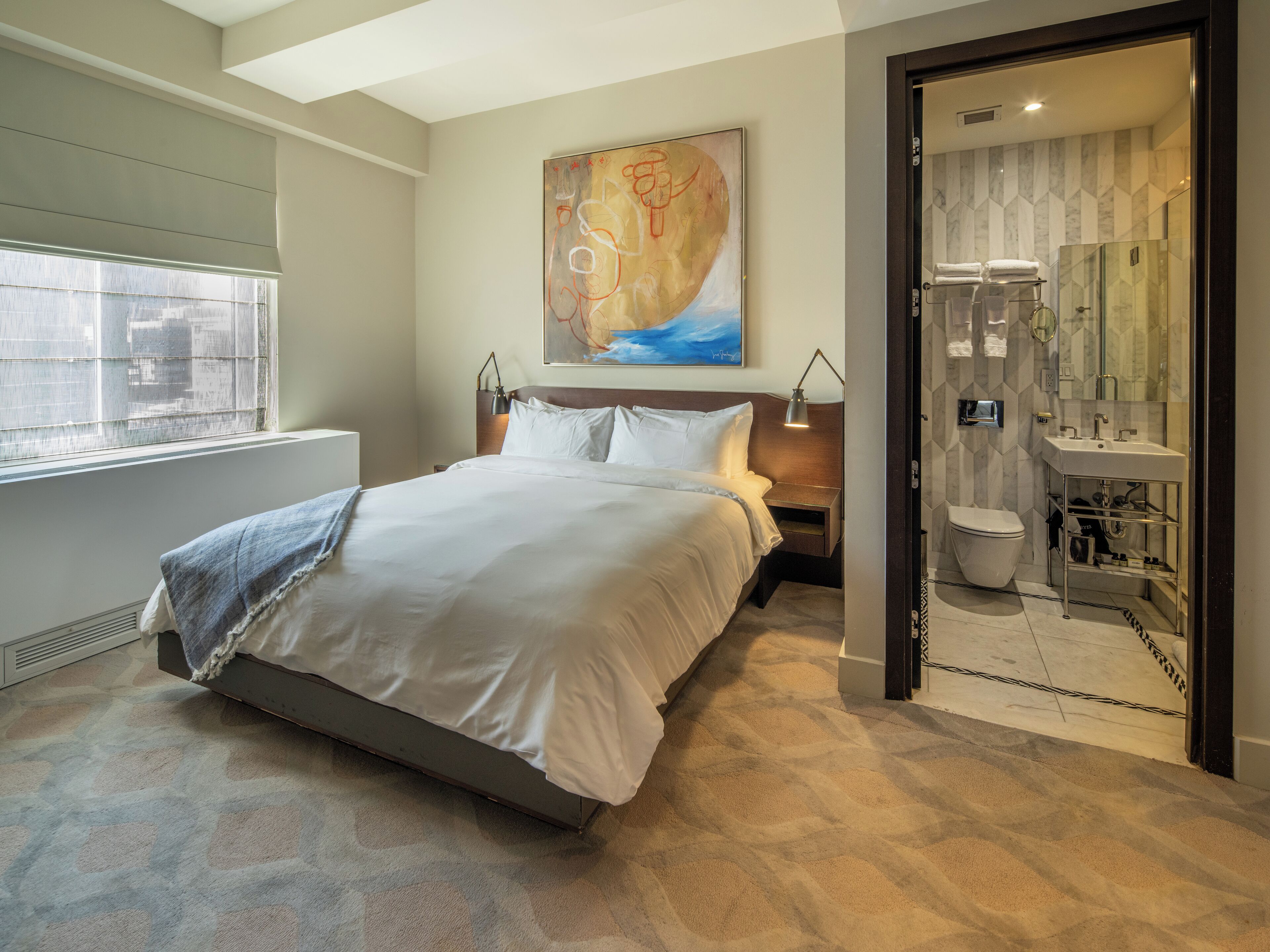 Quarto de hotel moderno com uma cama grande, um quadro na parede, banheiro com chuveiro de vidro e vista da cidade pela janela.