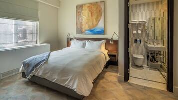 Quarto de hotel moderno com uma cama grande, um quadro na parede, banheiro com chuveiro de vidro e vista da cidade pela janela.