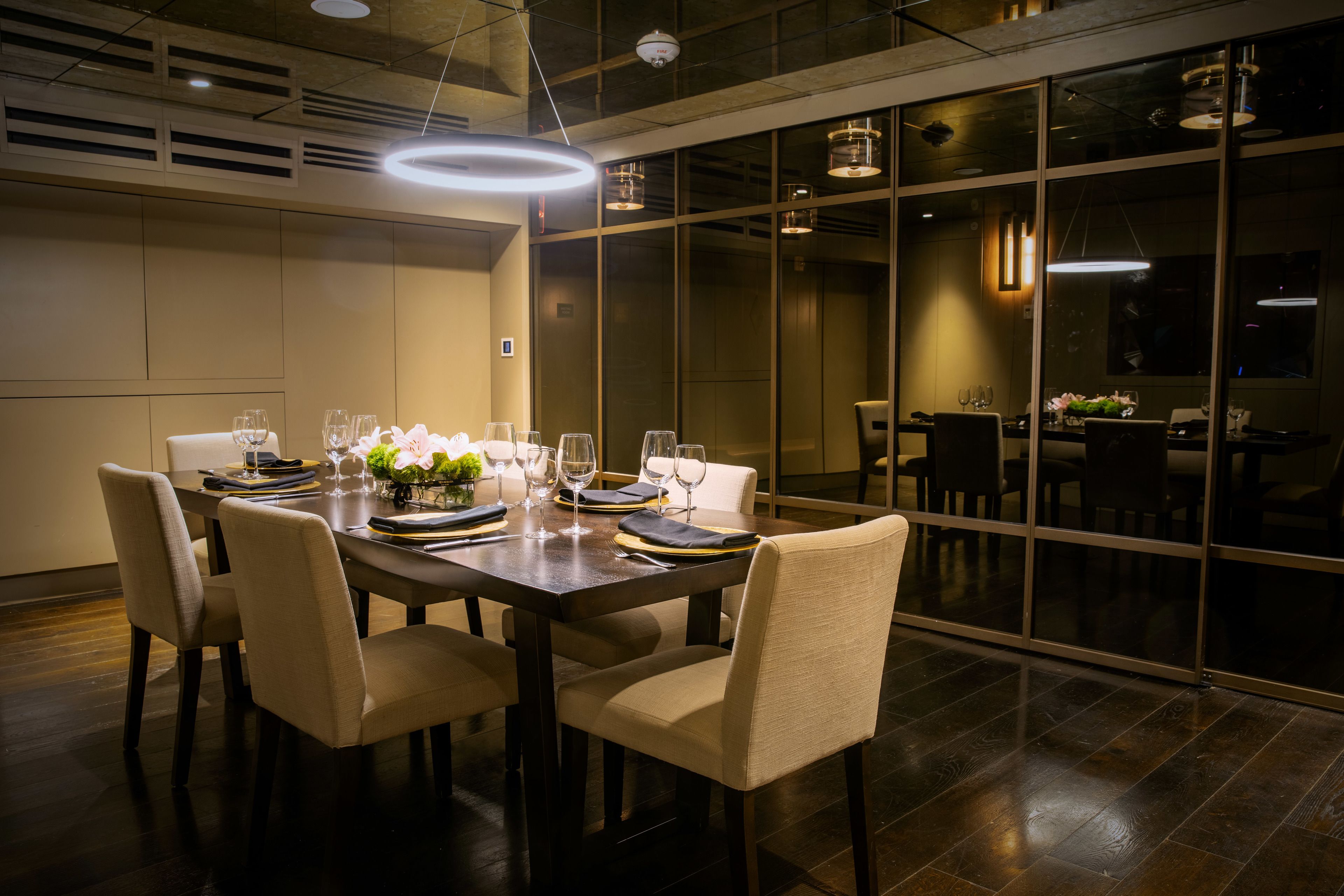 A dining area with a long table set for six, surrounded by chairs, with a view through large glass doors to another dining area.